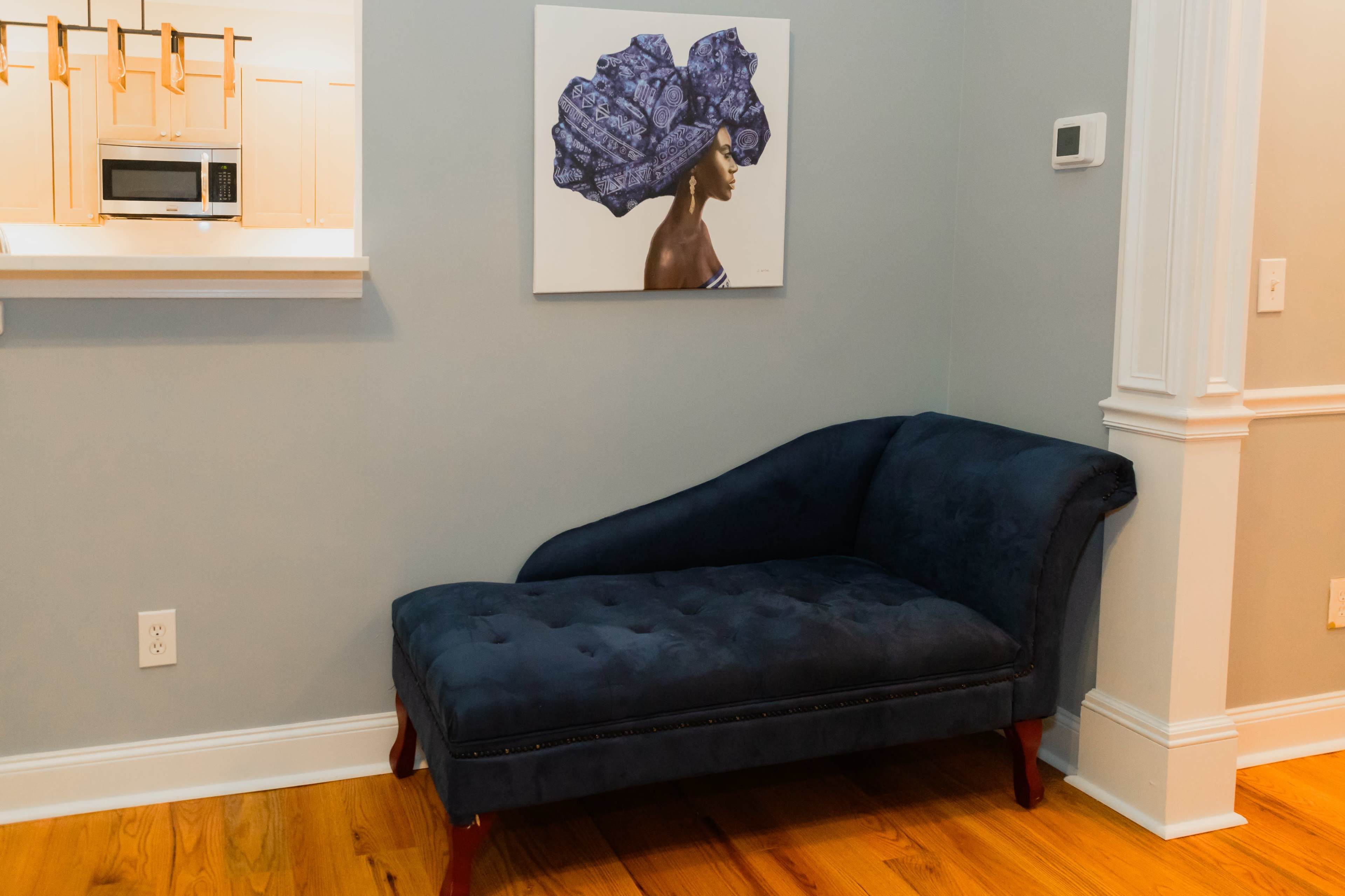 A dark blue chaise lounge is positioned against a gray wall, beneath a large artwork depicting a woman with a blue headwrap.