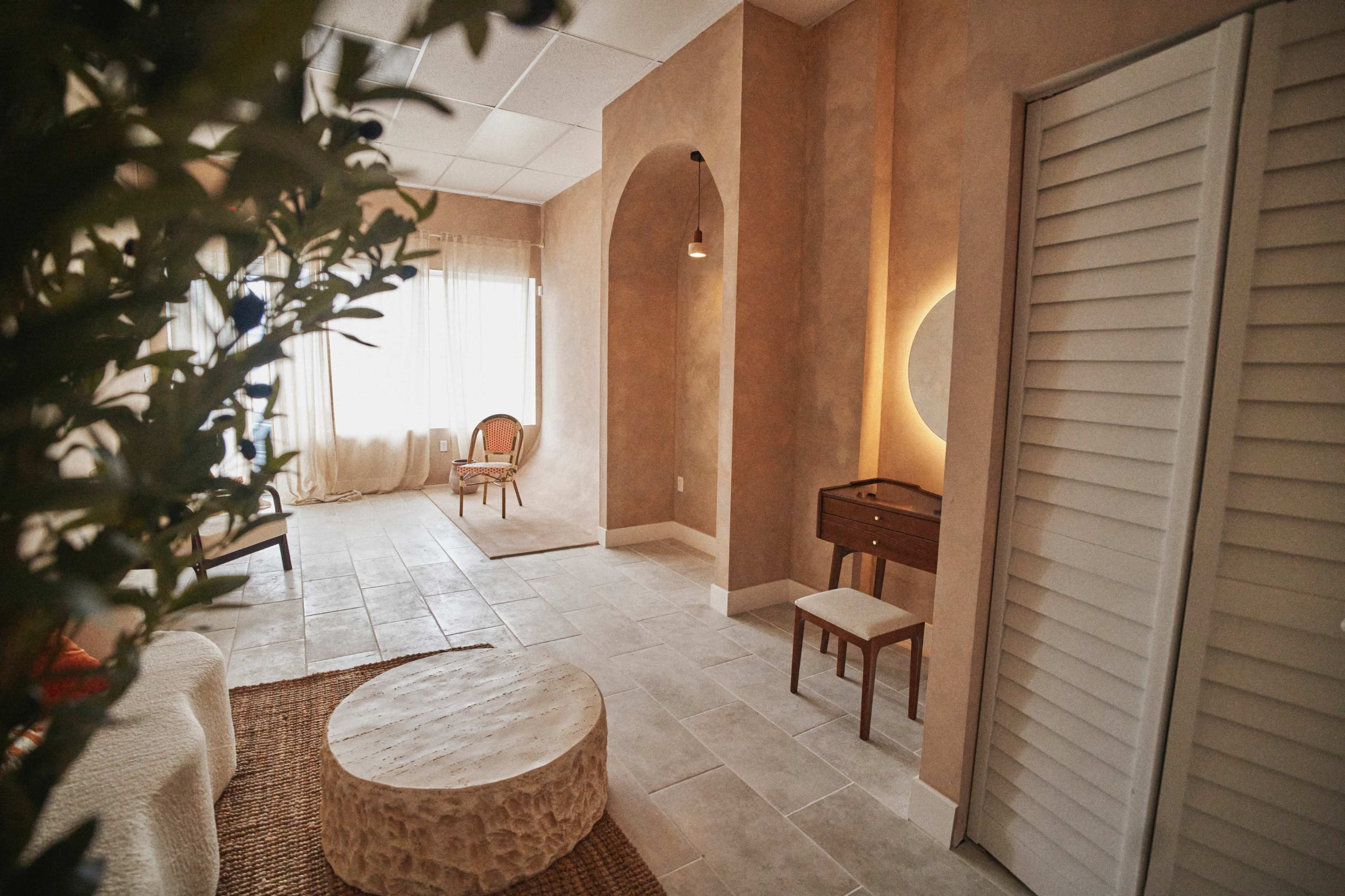 The image shows a softly lit interior space with neutral-colored walls, a round stone coffee table, a minimalist wooden chair, and a small dresser beneath a circular mirror.