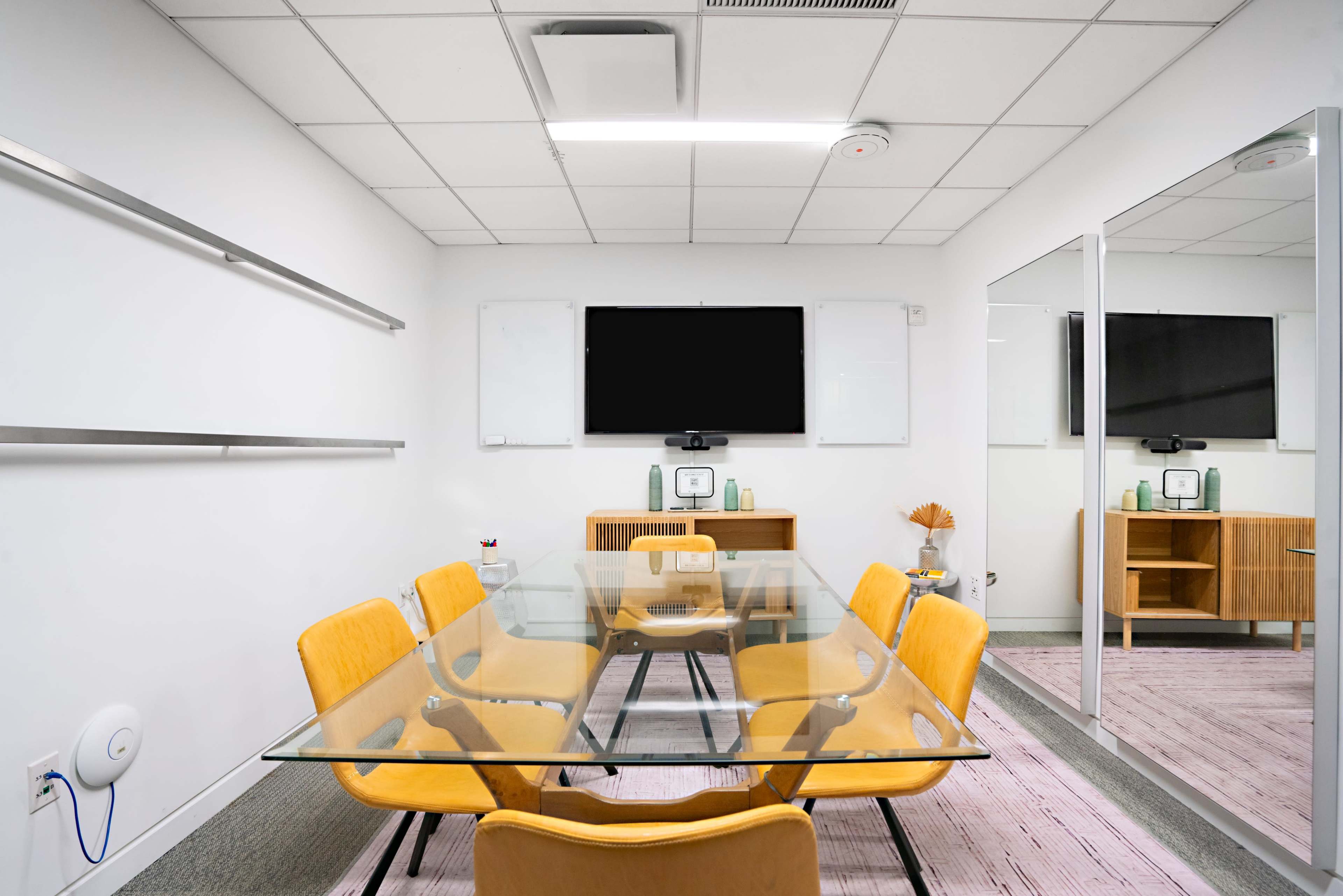 The image shows a modern conference room featuring a glass table surrounded by yellow chairs, a wall-mounted television, and a wooden cabinet.