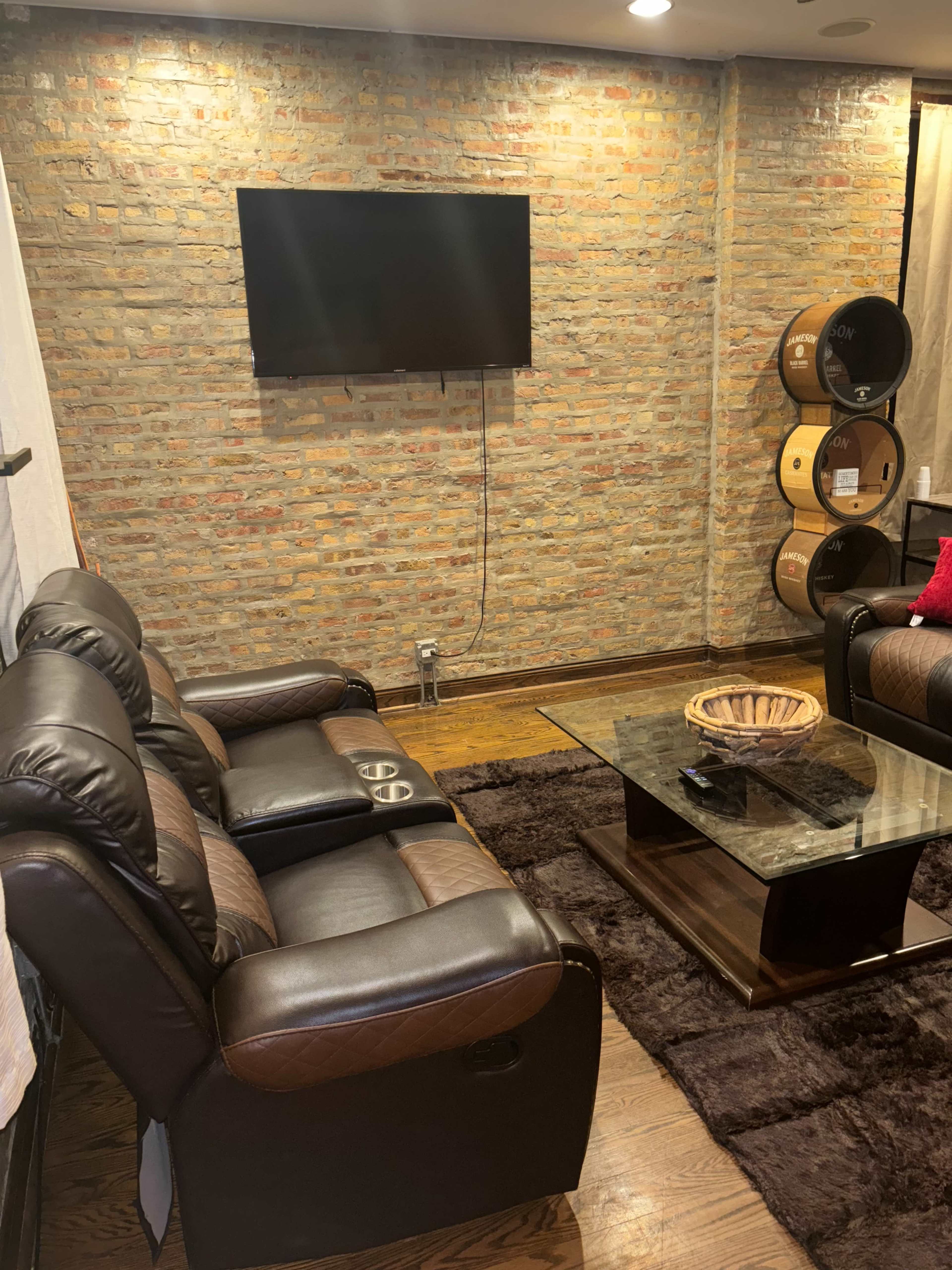 The image shows a cozy living room with a brick wall, two reclining sofas, a glass coffee table, and decorative wooden barrels in the background.