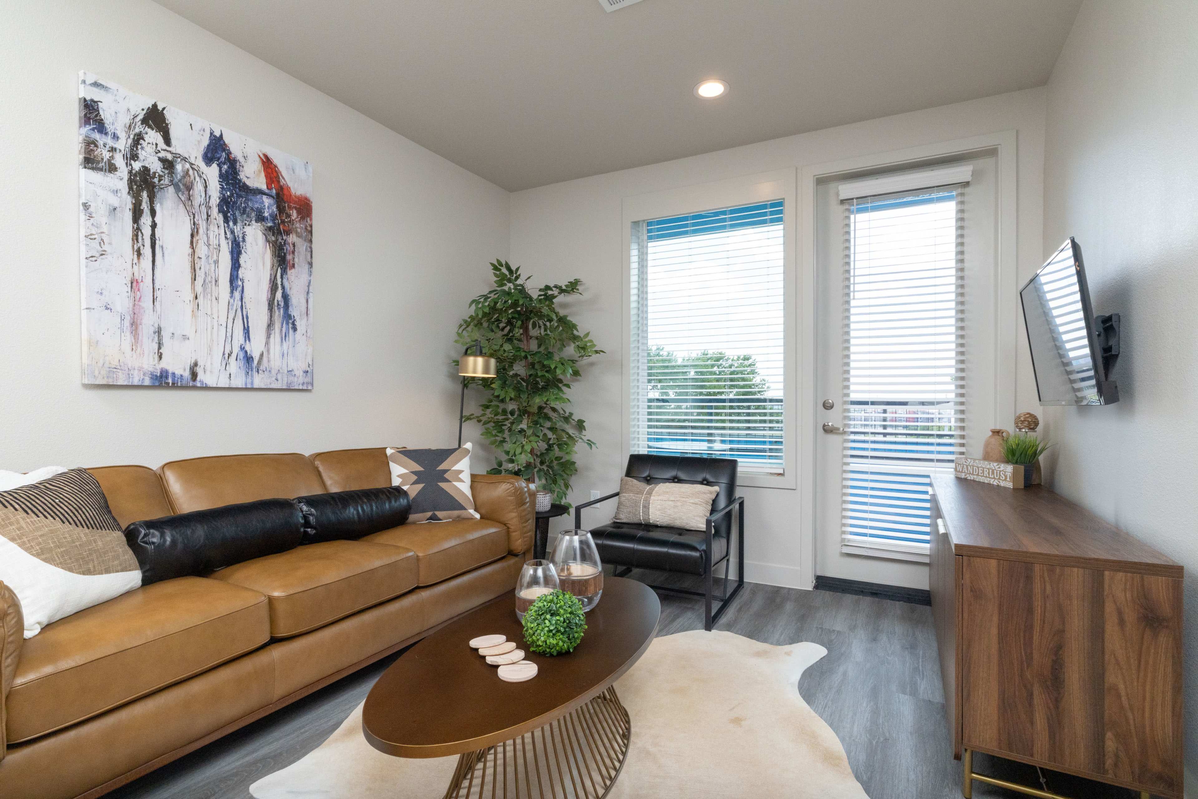 The image shows a modern living room featuring a brown leather sofa, a wooden TV stand, a small black chair, and a large artwork on the wall, with a window providing natural light.