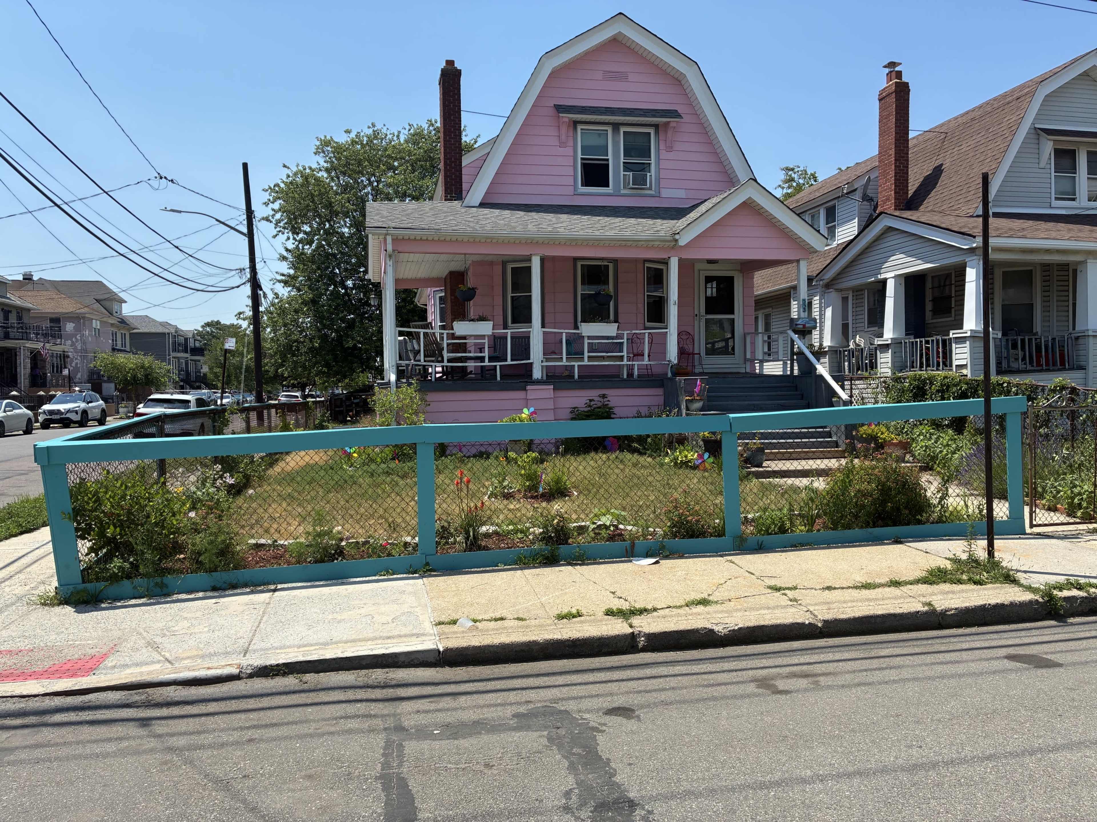 A pink house with a gabled roof has a garden surrounded by a teal fence in front of it.