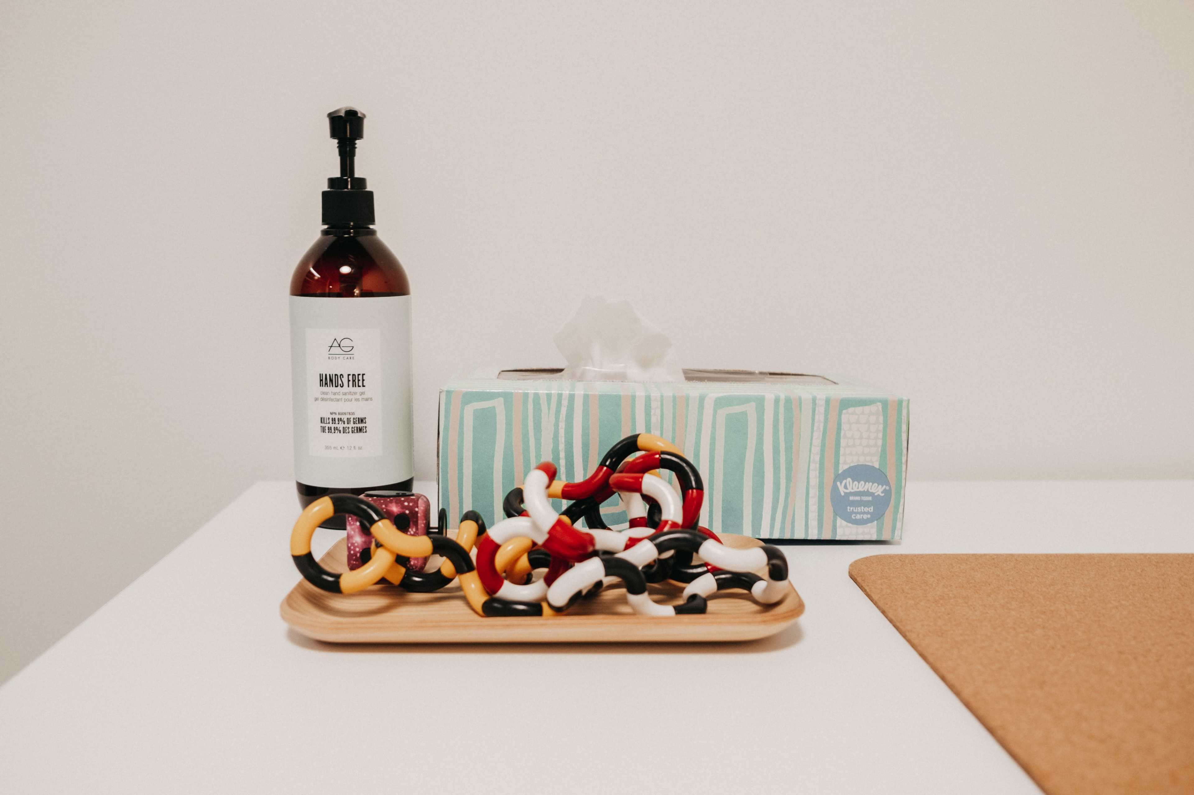 A bottle of hand sanitizer, a tissue box, and a tray of colorful hair ties are arranged on a white table.