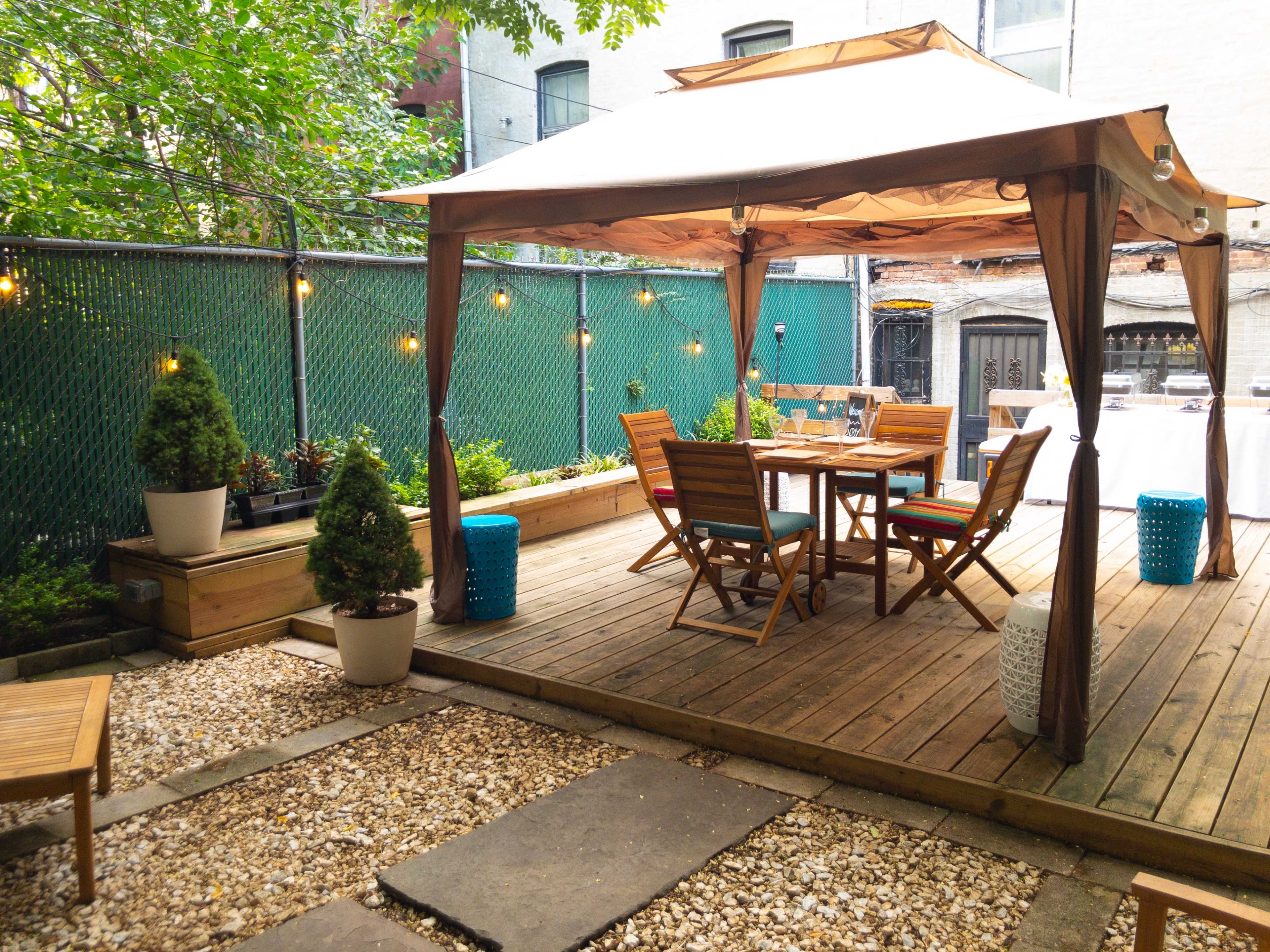 A wooden deck with a gazebo, outdoor dining furniture, and decorative planters surrounded by greenery and a fence.