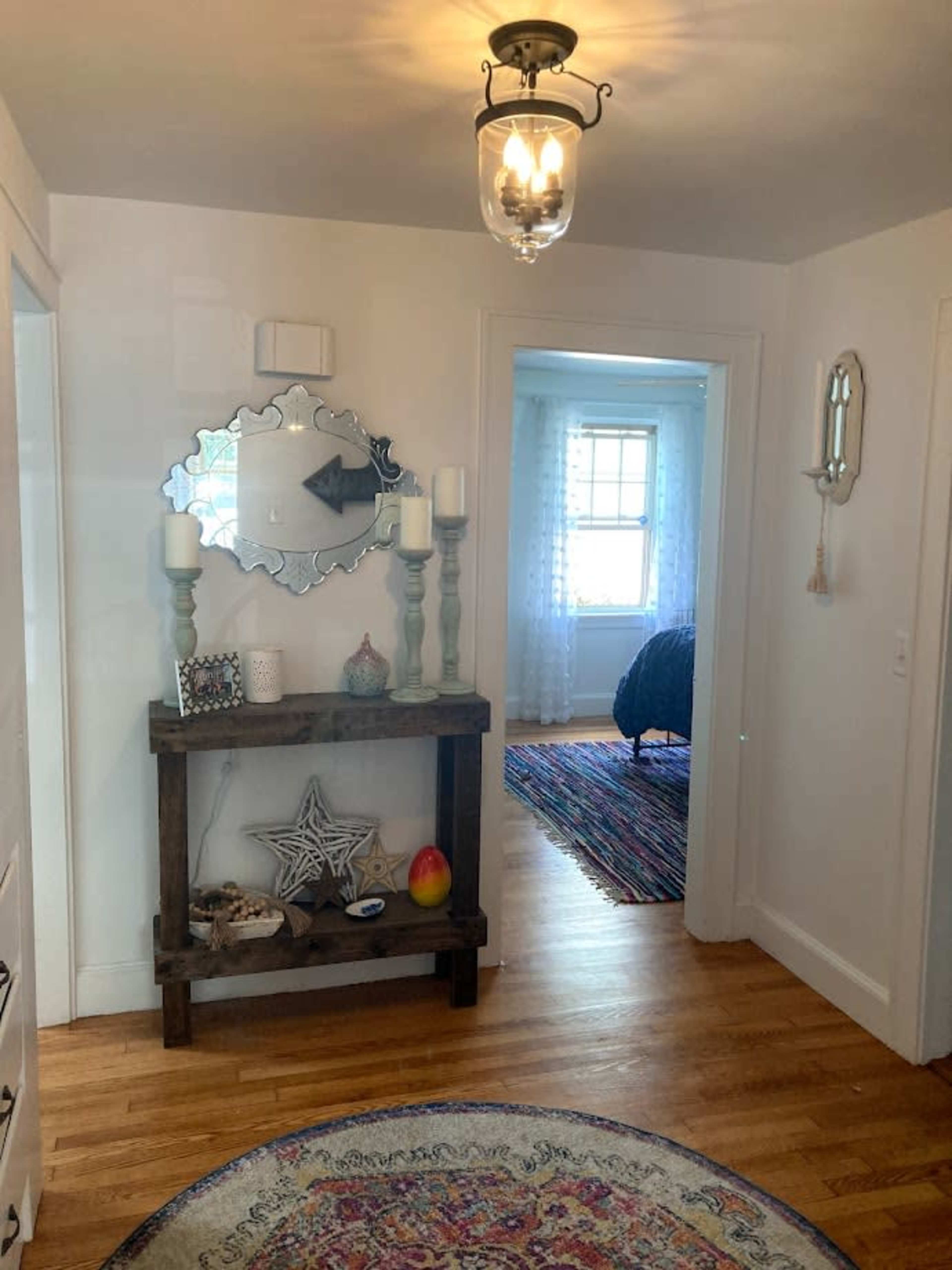 A bright hallway with a wooden console table displaying decorative items and a mirror, leading to a room with a bed and colorful rug.