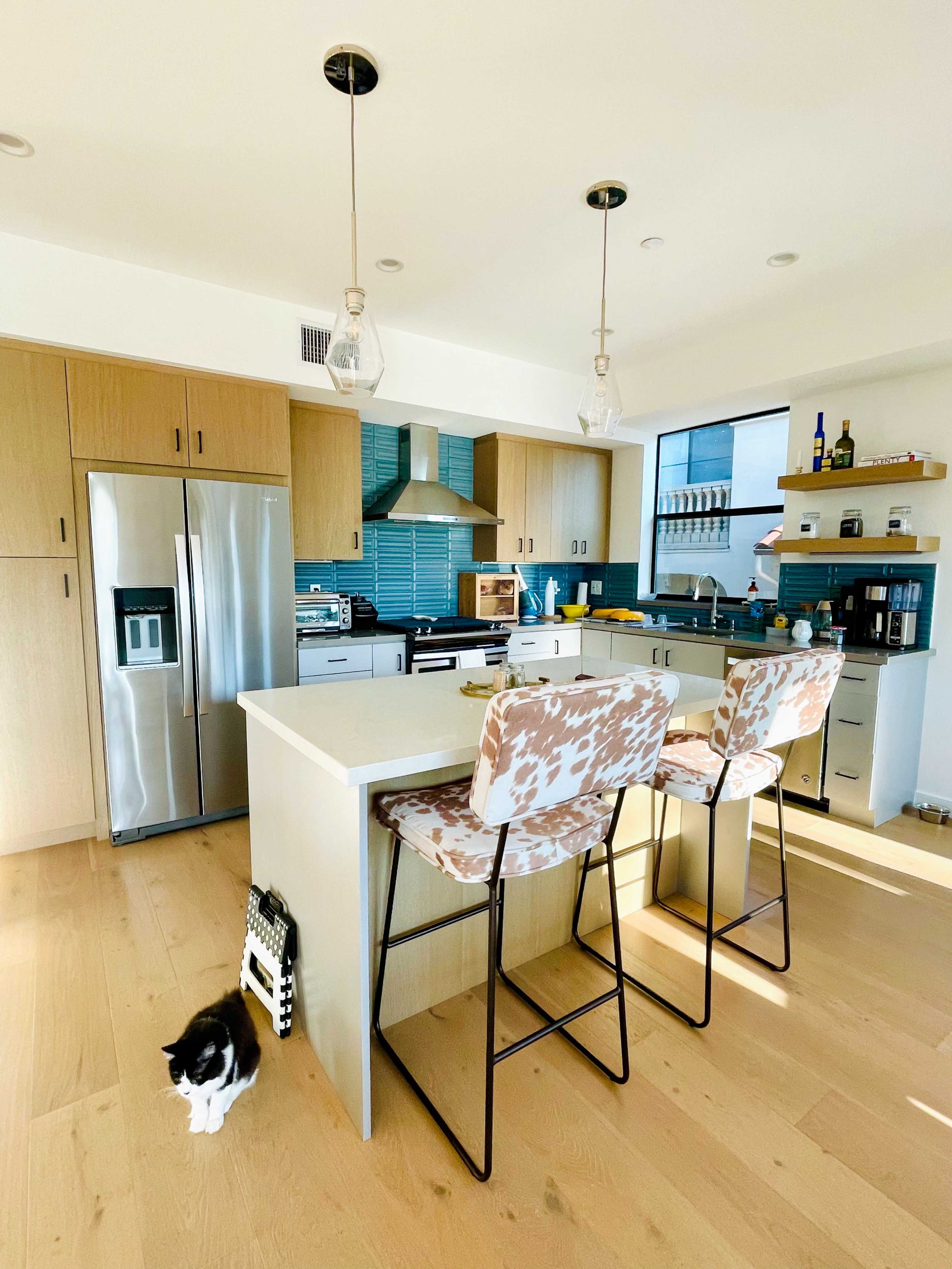 A modern kitchen features stainless steel appliances, a central island with two patterned stools, and a cat resting on the wooden floor.