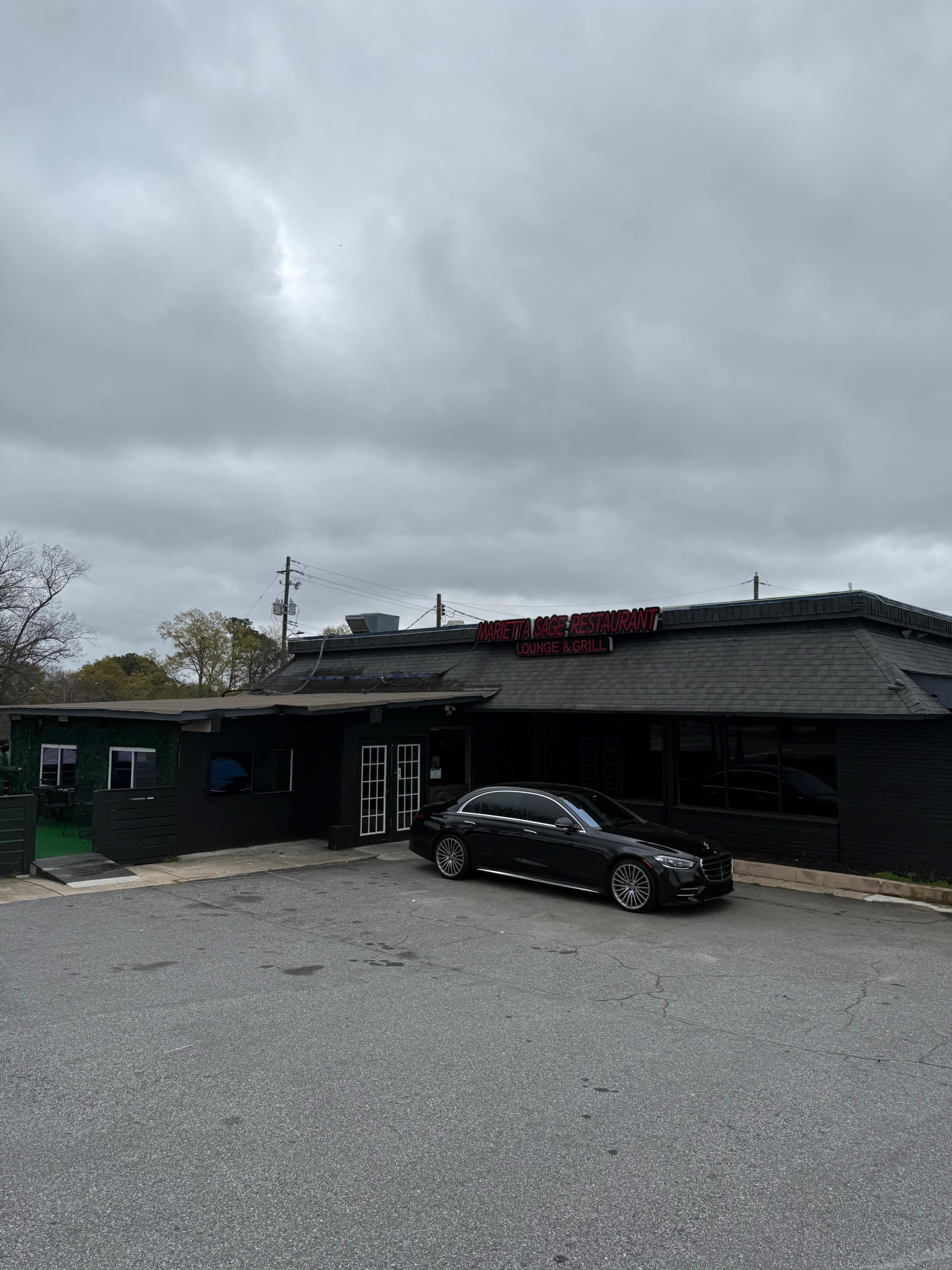 The image shows a dark-colored building with a signage indicating it is a restaurant, accompanied by a parked black car in front, under a cloudy sky.