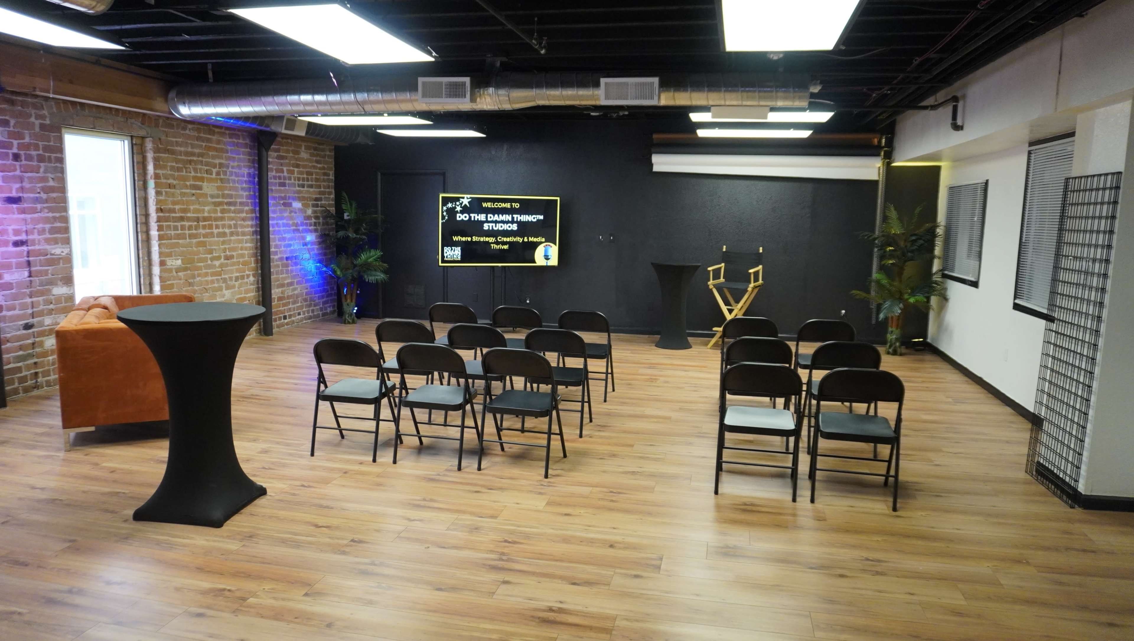 A modern event space with rows of black chairs facing a screen, a high table, and a director's chair, all set against brick and black walls.