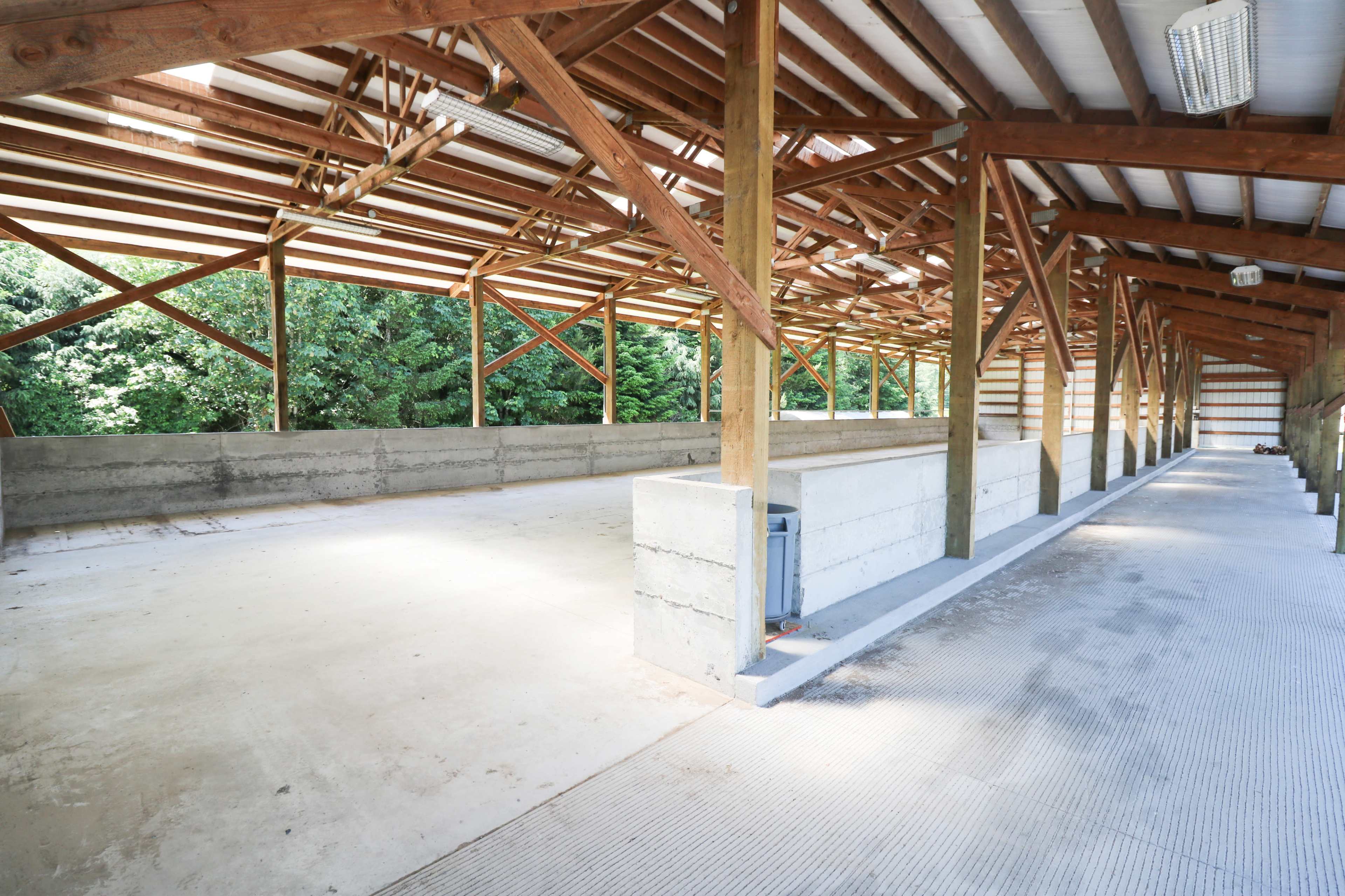 The image shows an empty barn with a concrete floor, wooden beams, and open sides facing a tree line.