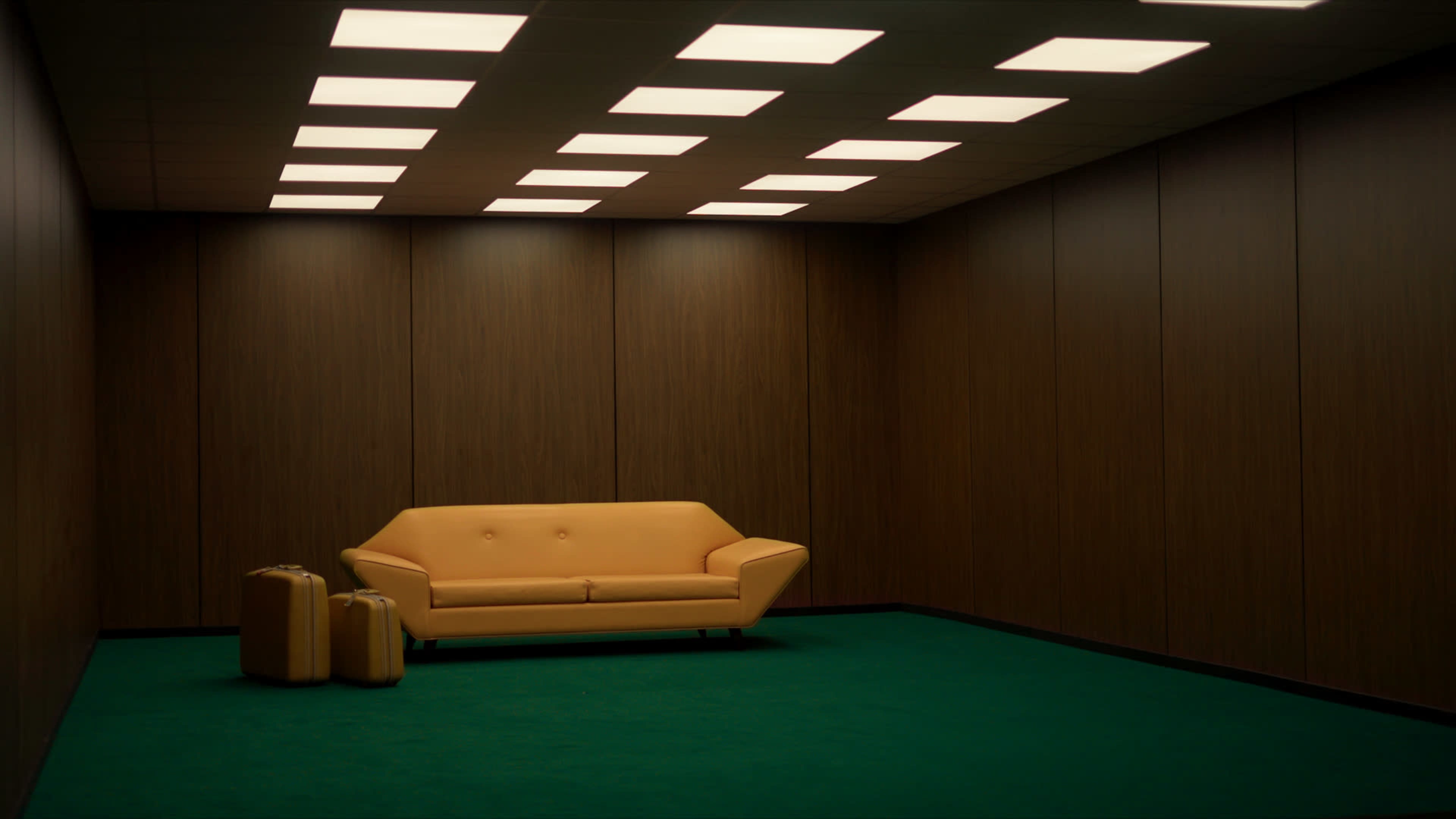 The image shows a sparsely furnished room with brown paneling, a yellow couch, and two yellow suitcases on a green carpet.