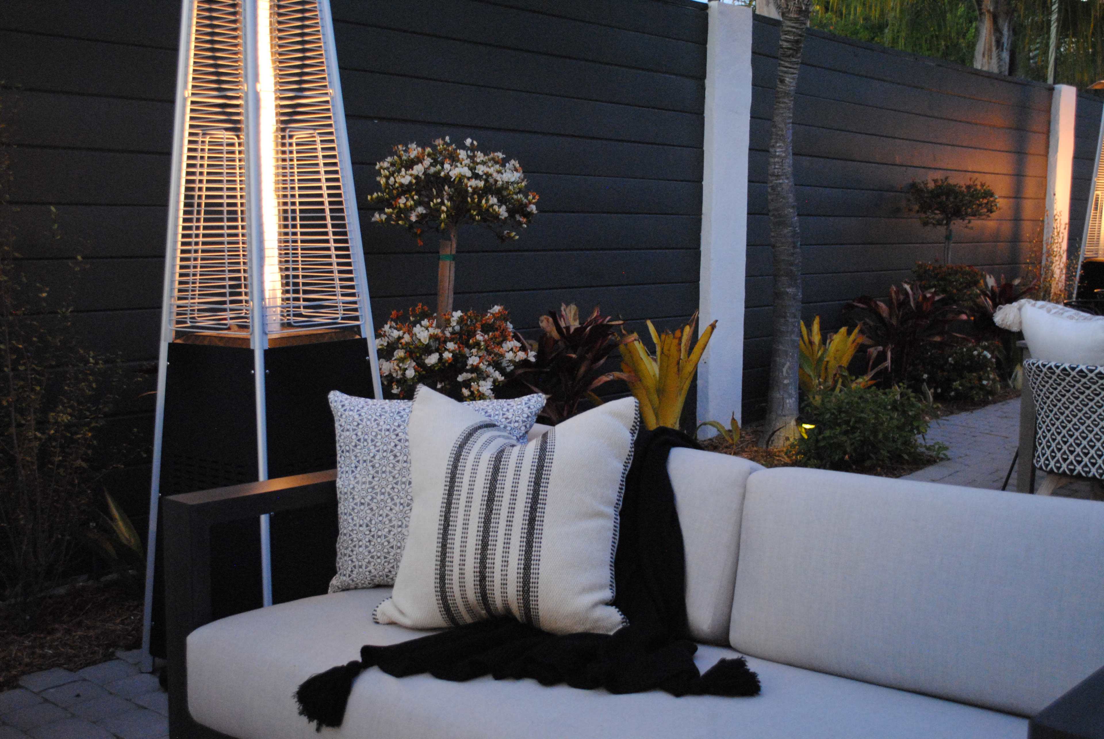 A modern outdoor seating area features a lighted heater beside a sofa adorned with decorative pillows and a throw blanket, surrounded by landscaped plants and flowers.