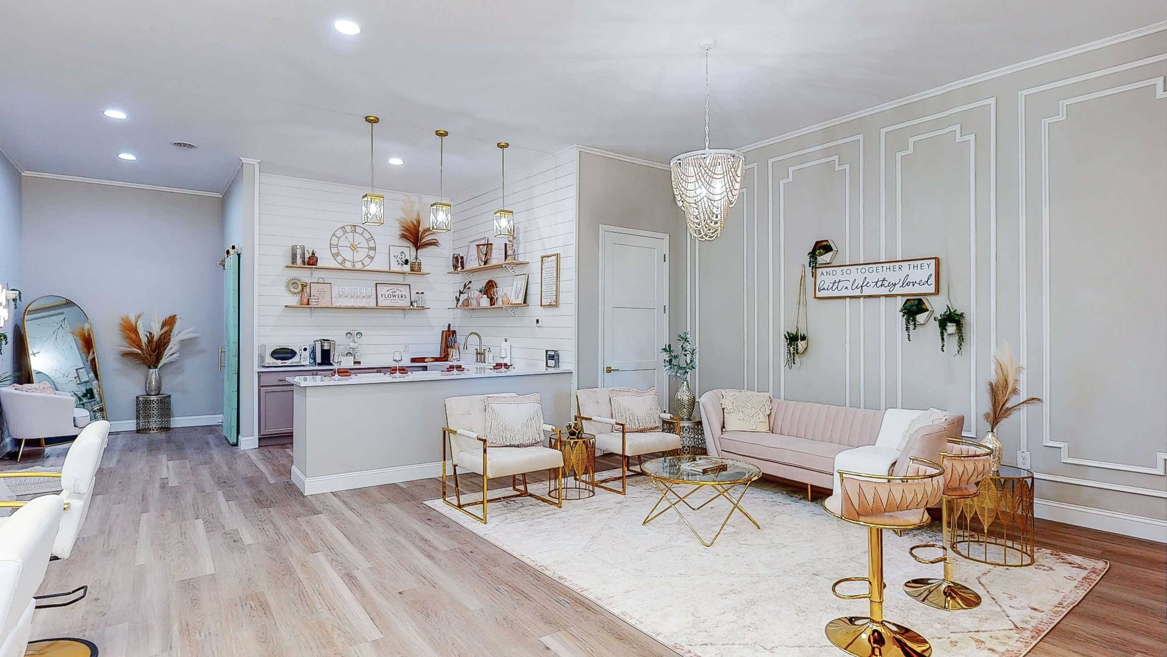 A modern, stylish living space featuring a pink sofa, elegant light fixtures, a rug, and a kitchenette with decorative wall shelves.