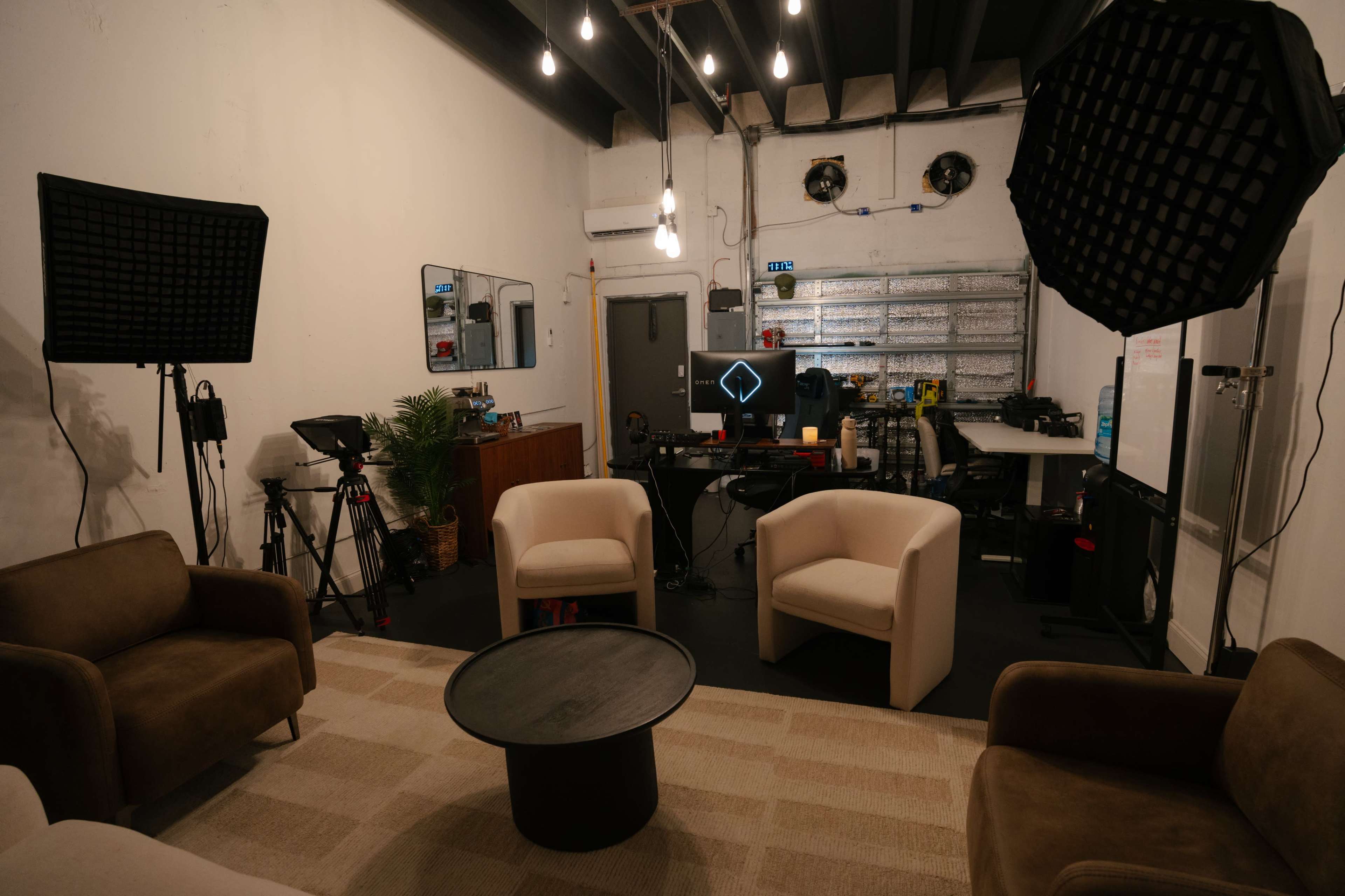 The image shows a modern studio space with a seating area, two soft chairs, a coffee table, and various filming equipment arranged around a computer workspace.