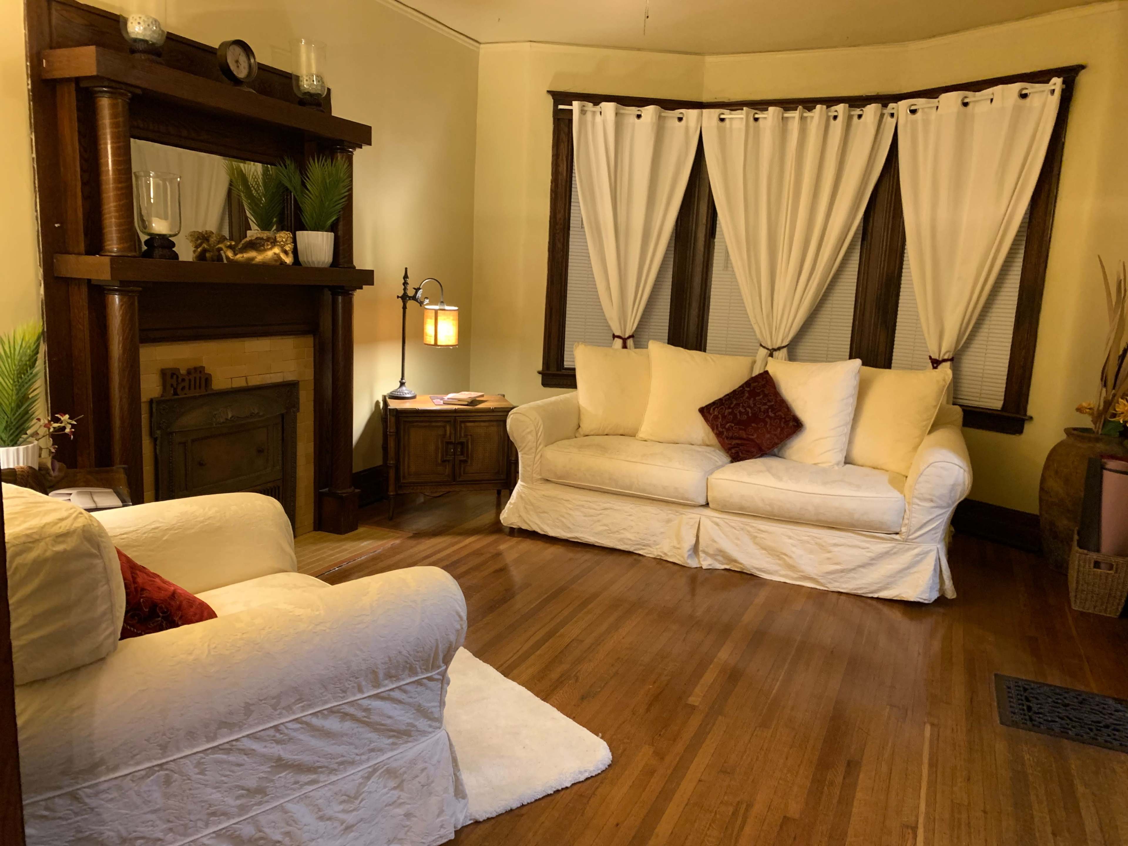 The image shows a cozy living room with two white sofas, a wooden bookshelf, and large windows draped with white curtains.