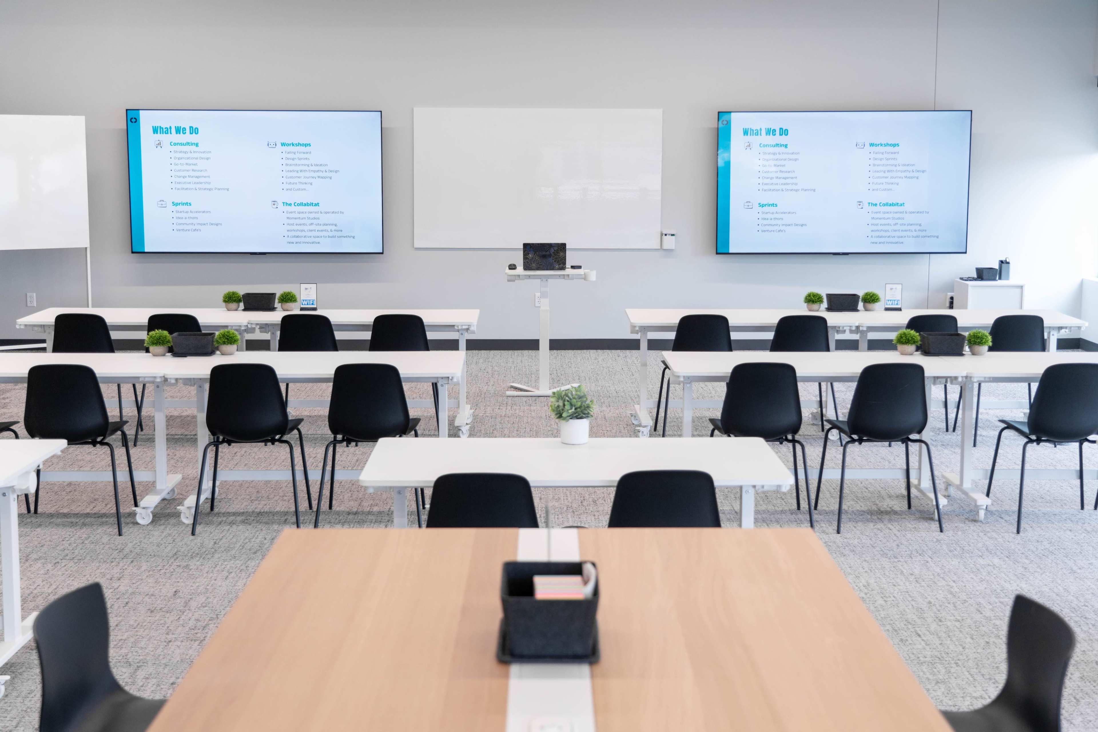 The image shows a modern classroom setup with rows of chairs and tables facing two large screens displaying information, and a minimalistic design with potted plants.
