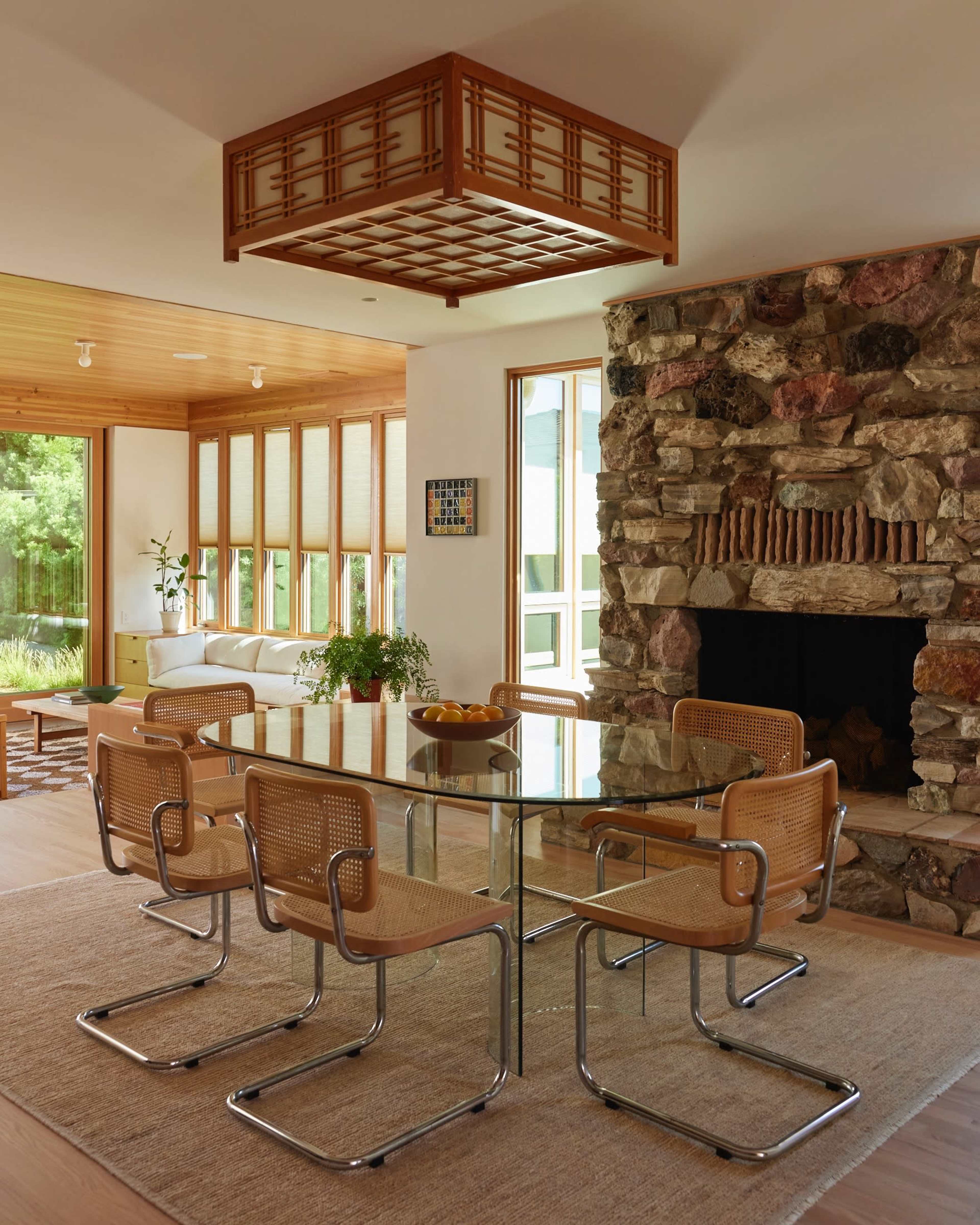 A modern dining area features a glass table surrounded by six rattan chairs, with a stone fireplace and large windows in the background.