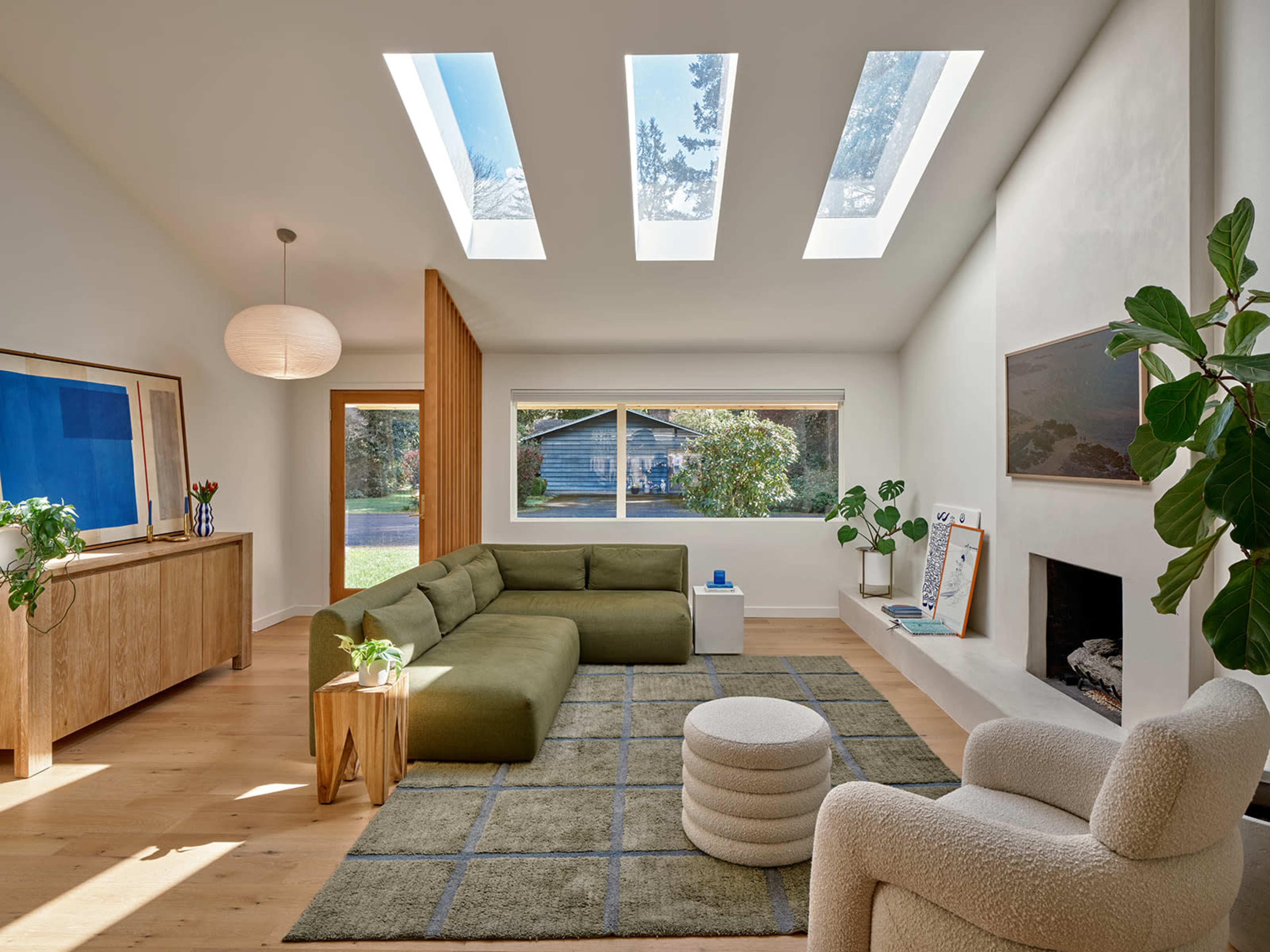 The image depicts a modern living room with a green sectional sofa, a round white ottoman, and large skylights allowing natural light to fill the space.