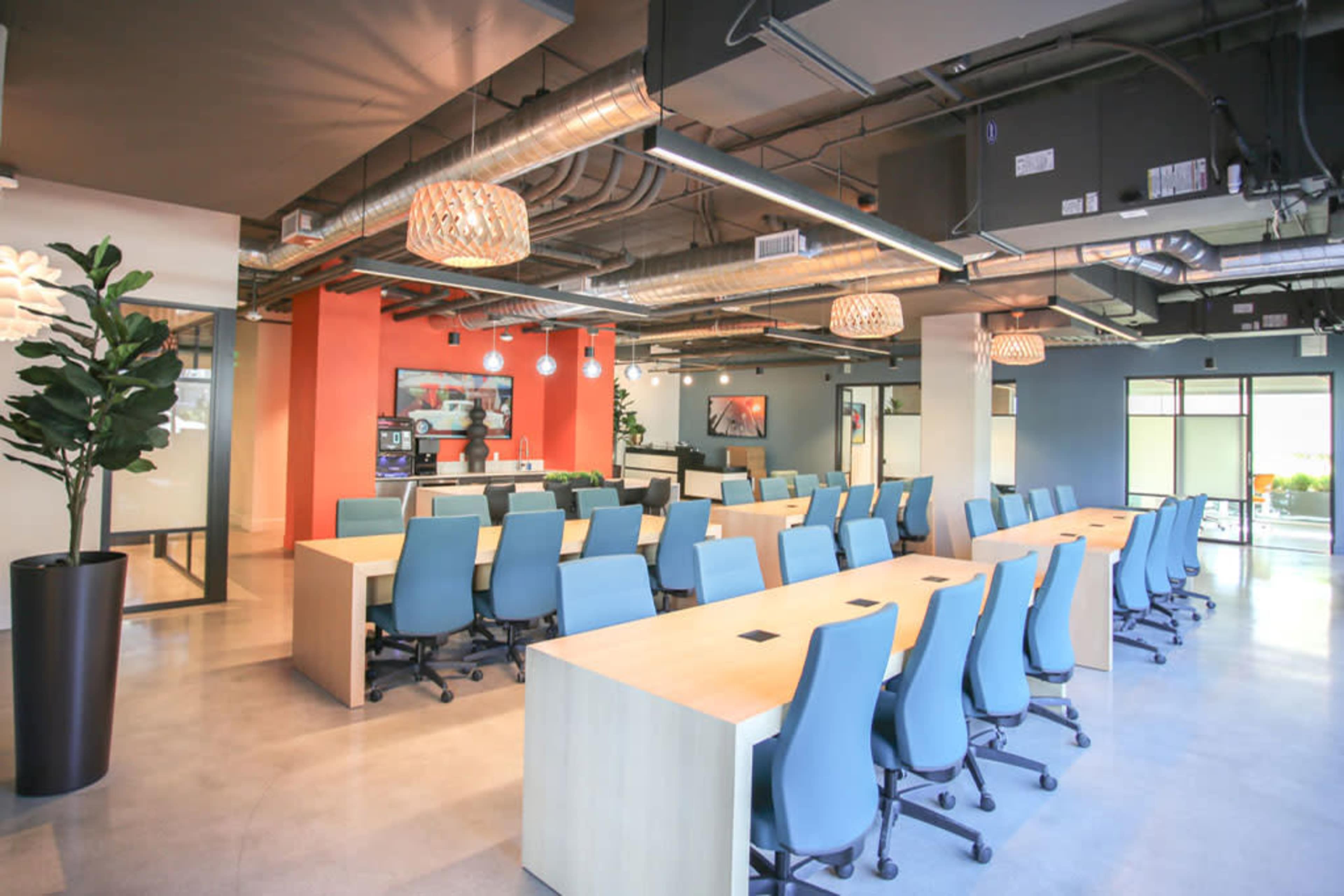 A modern office space with a long communal table surrounded by blue chairs, brightly colored walls, and hanging light fixtures.
