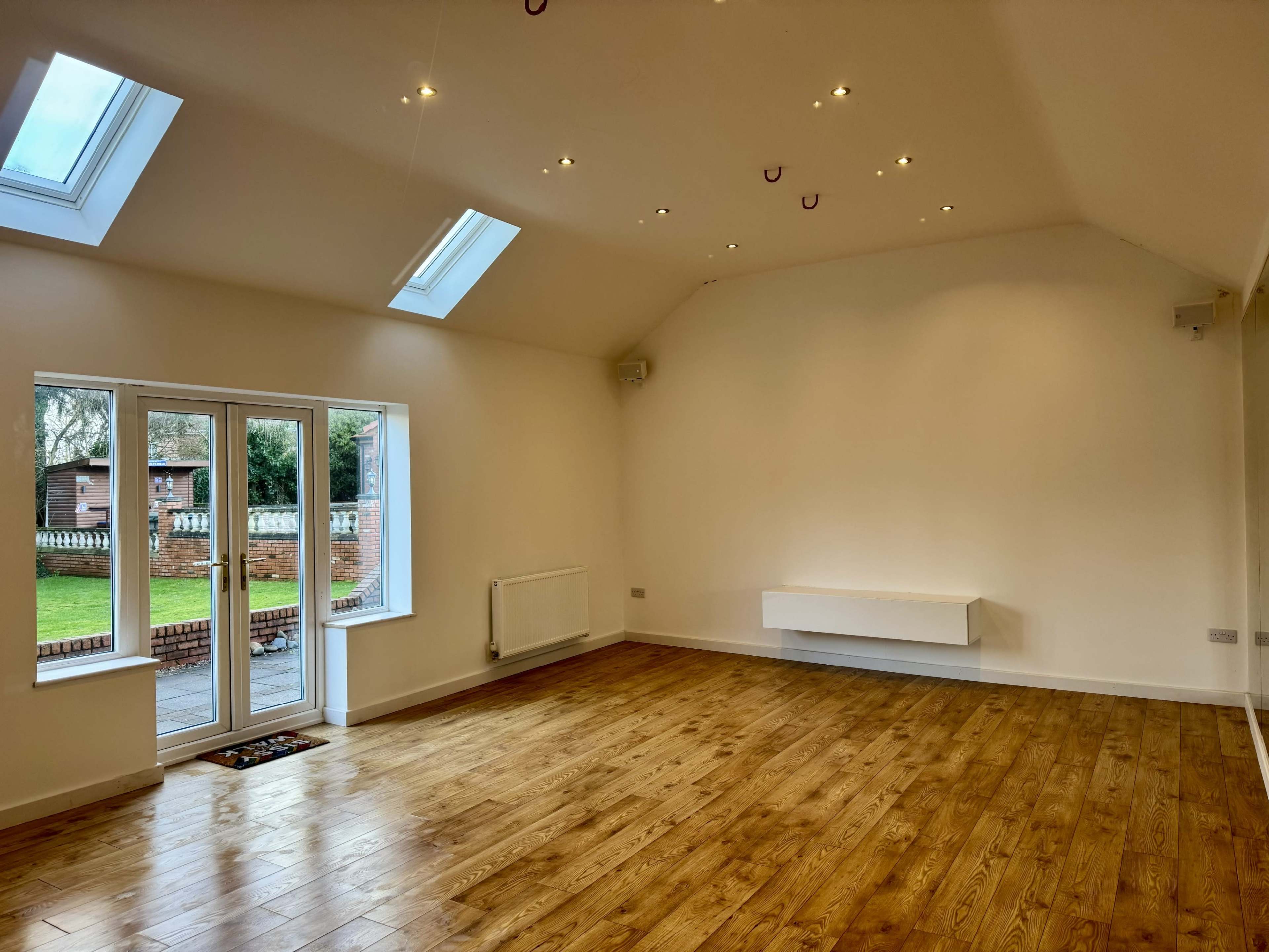 The image shows a spacious, empty room with wooden flooring, large windows, and a high ceiling illuminated by recessed lighting.