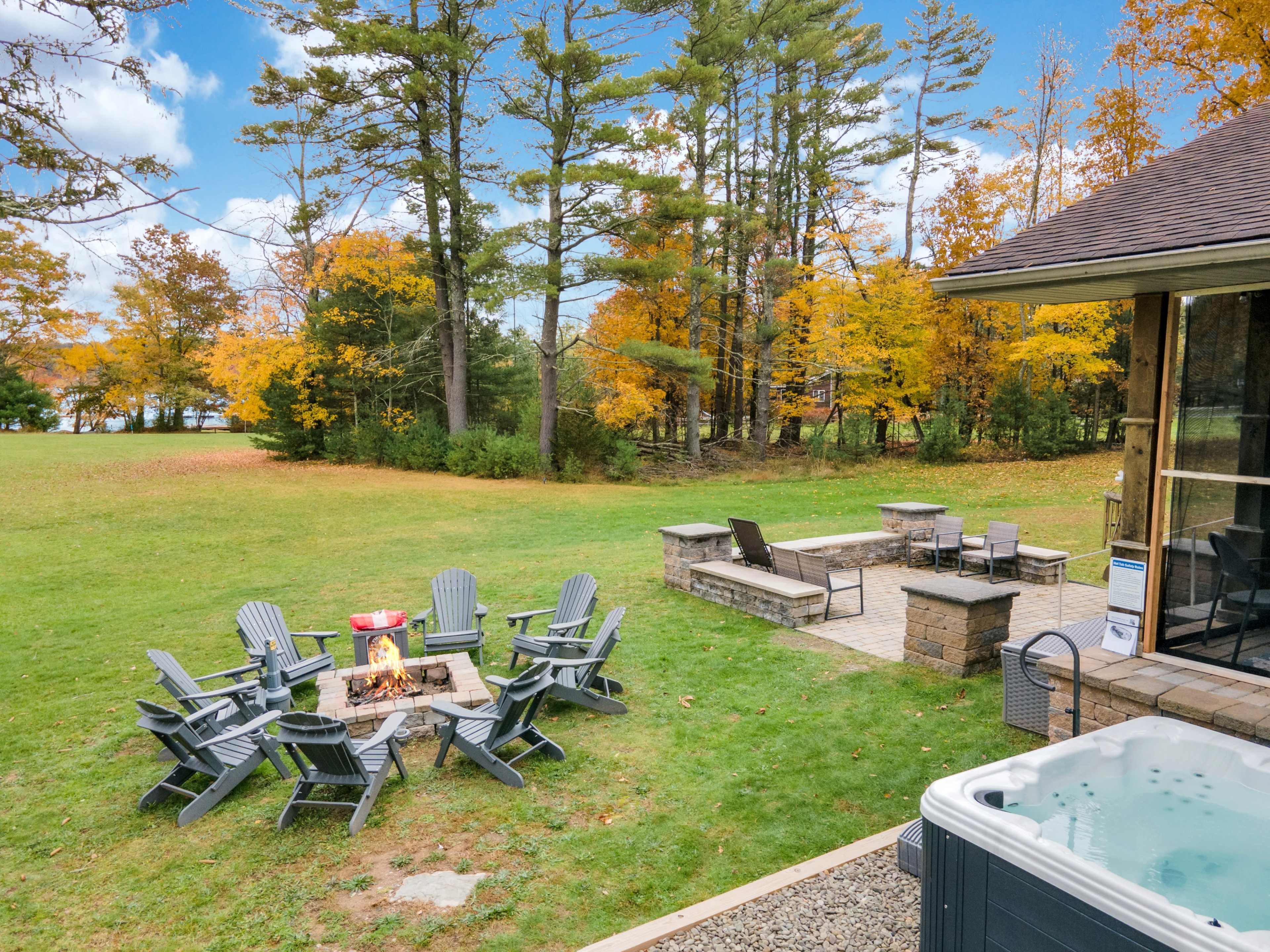 A fire pit surrounded by adirondack chairs is set in a grassy area near a hot tub, with trees displaying autumn foliage in the background.