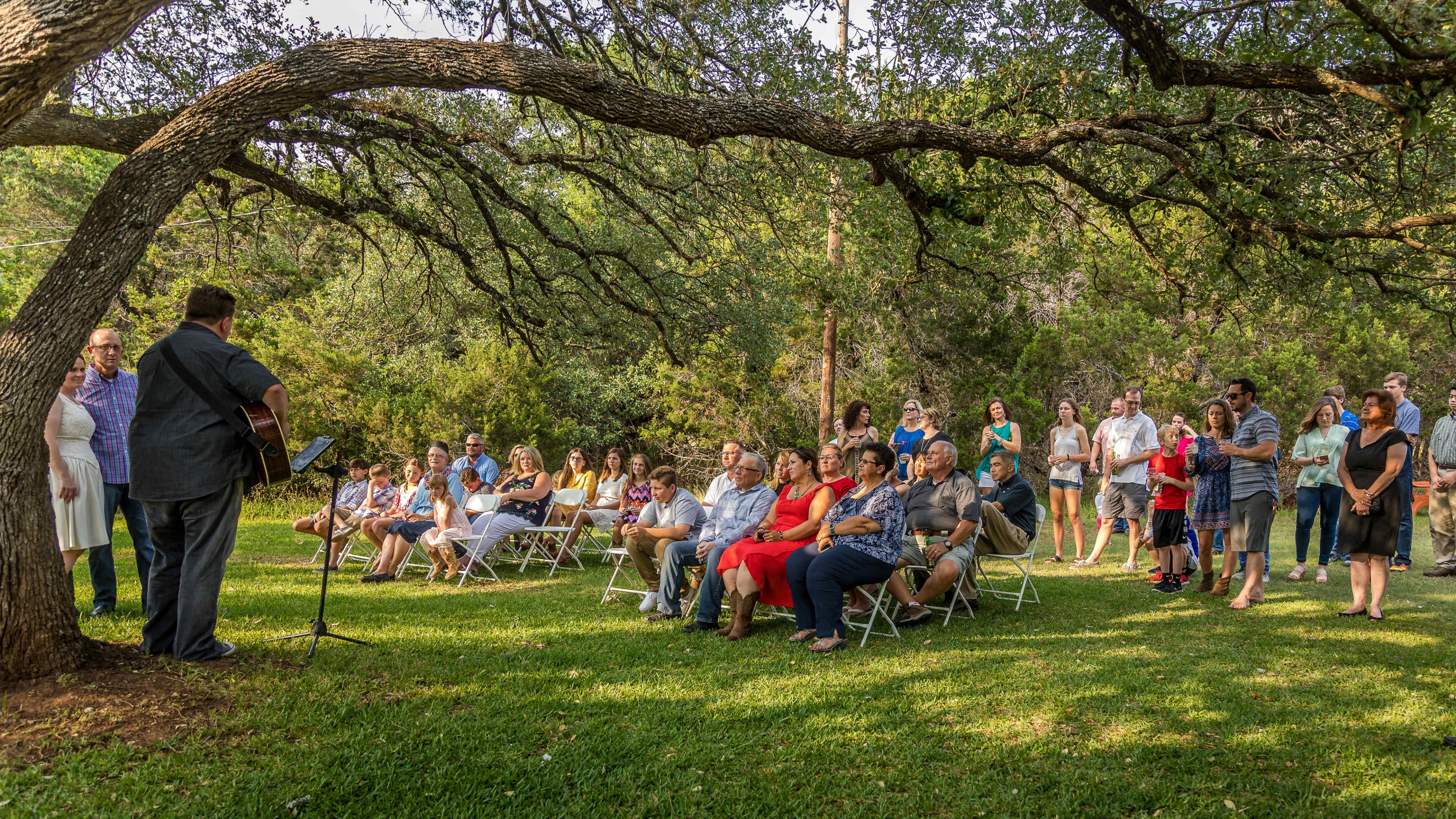 A musician performs on a grassy area under a large tree, while an audience sits in white chairs, listening attentively.