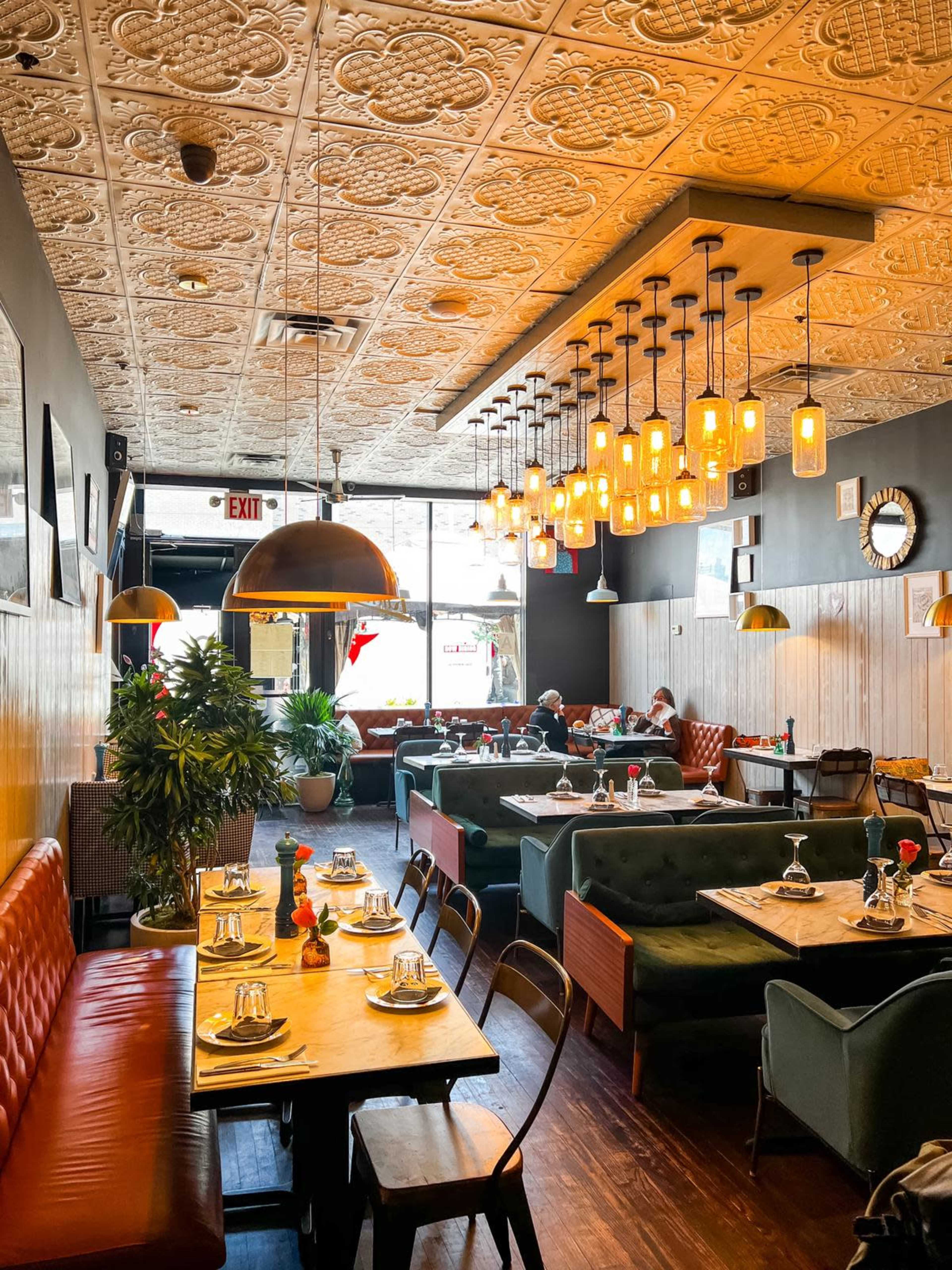 The image shows a modern restaurant interior with an ornate ceiling, wooden tables set for dining, and a mix of green and red seating arrangements.