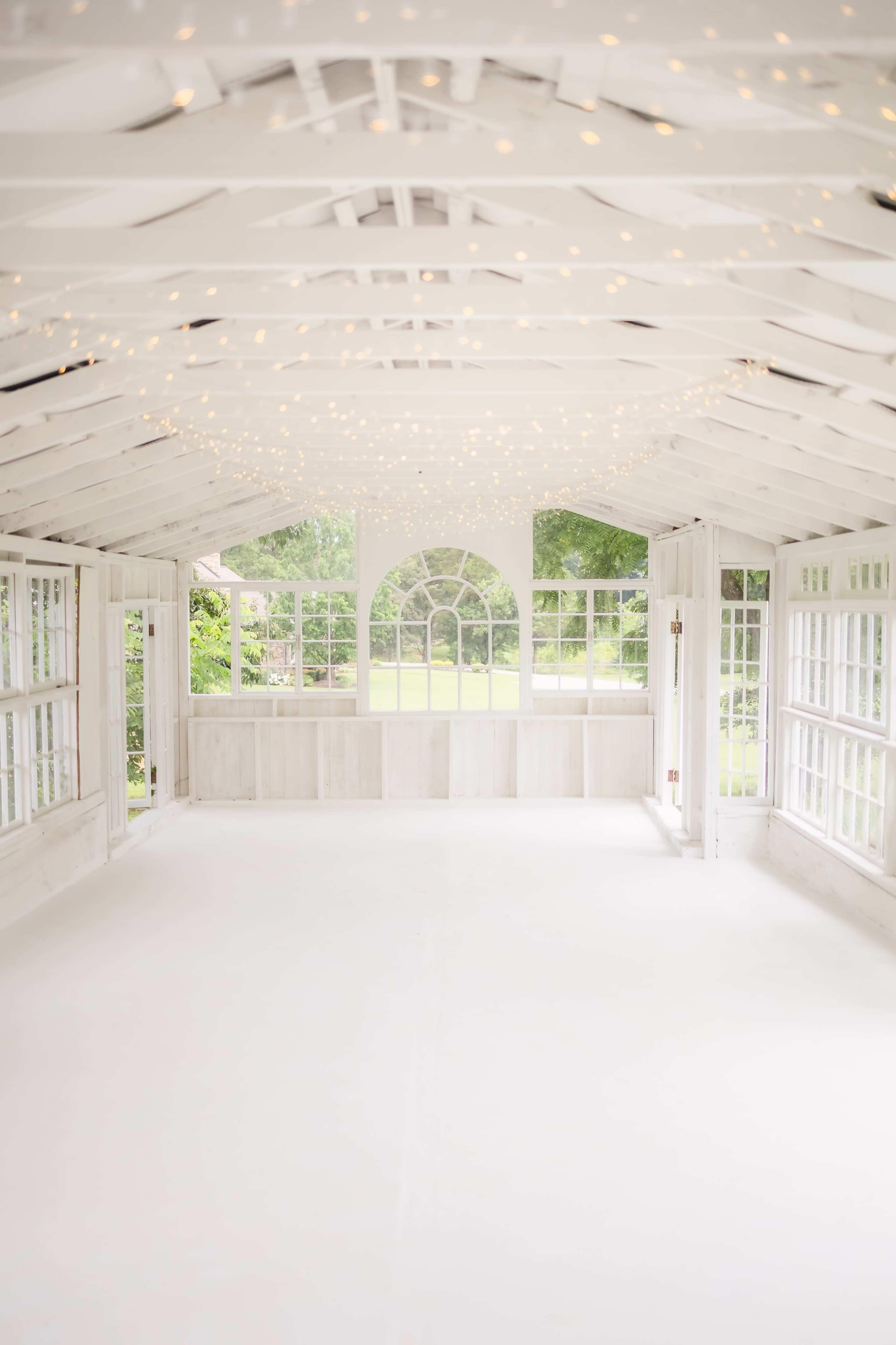 The image shows a spacious, well-lit room with white walls and wooden beams, featuring numerous windows that provide a view of greenery outside.