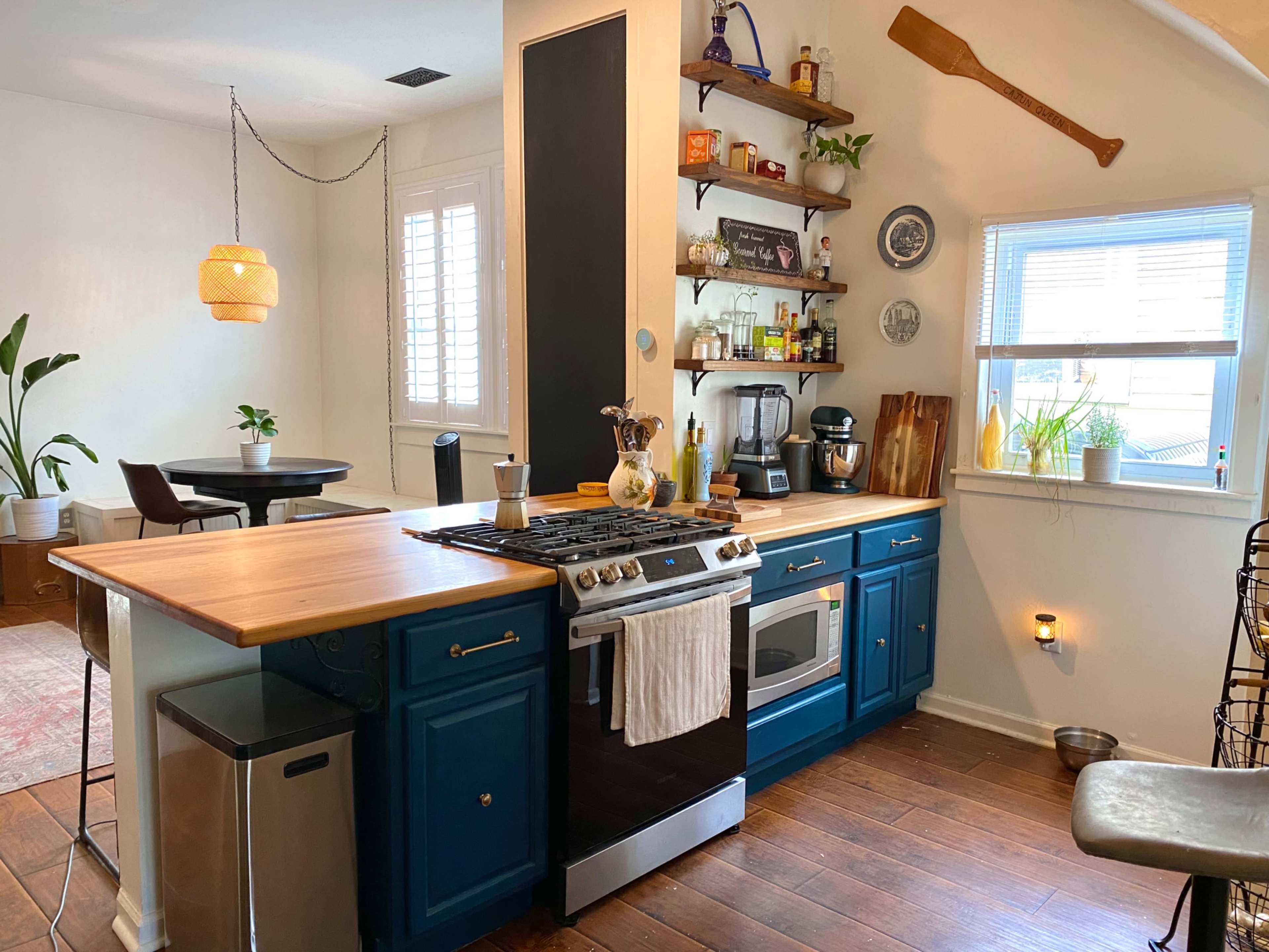 The image shows a modern kitchen with blue cabinetry, a wooden island, a stainless steel stove, and open shelving displaying various kitchen items.