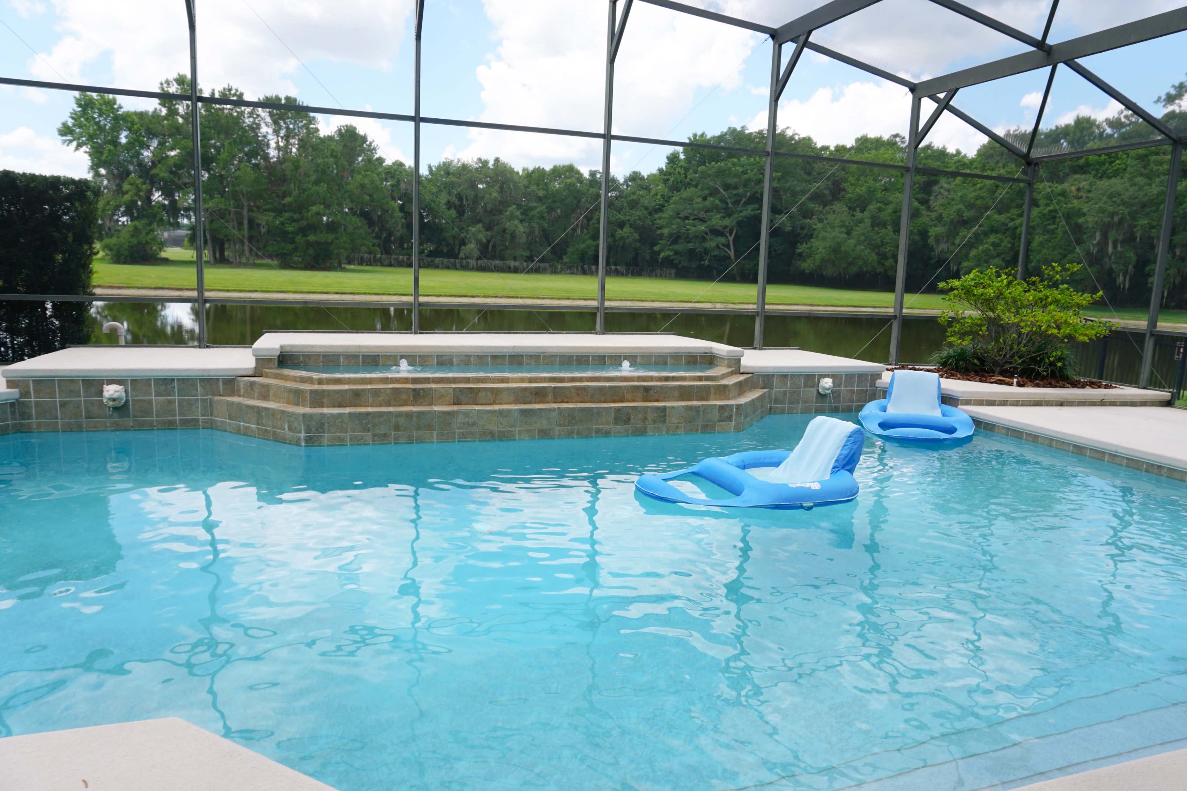 A swimming pool with two blue loungers, a raised spa area, and a view of a green landscape through a screened enclosure.