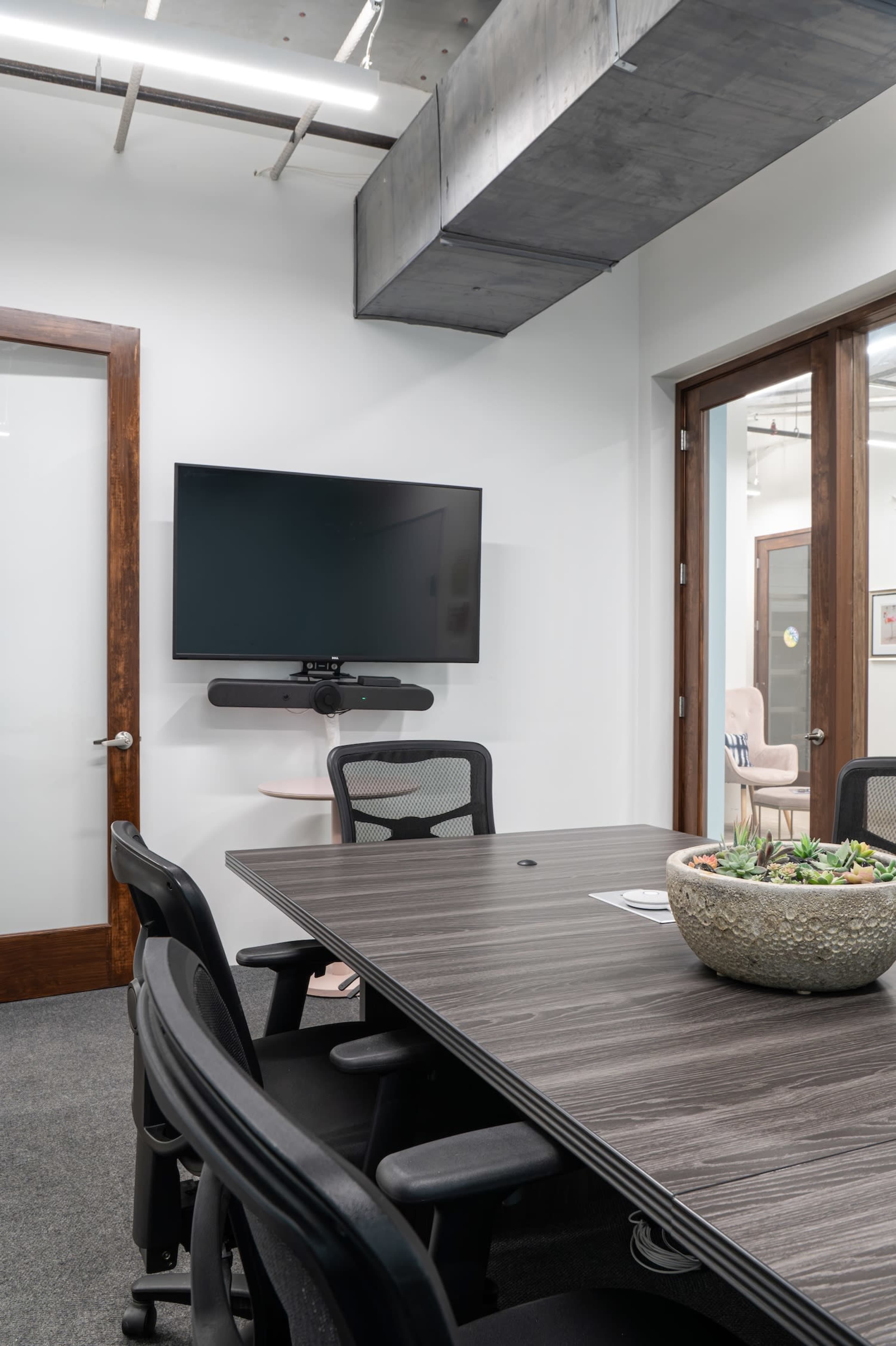 A modern conference room features a large table surrounded by black chairs, with a flat-screen TV mounted on the wall and a potted plant centerpiece.