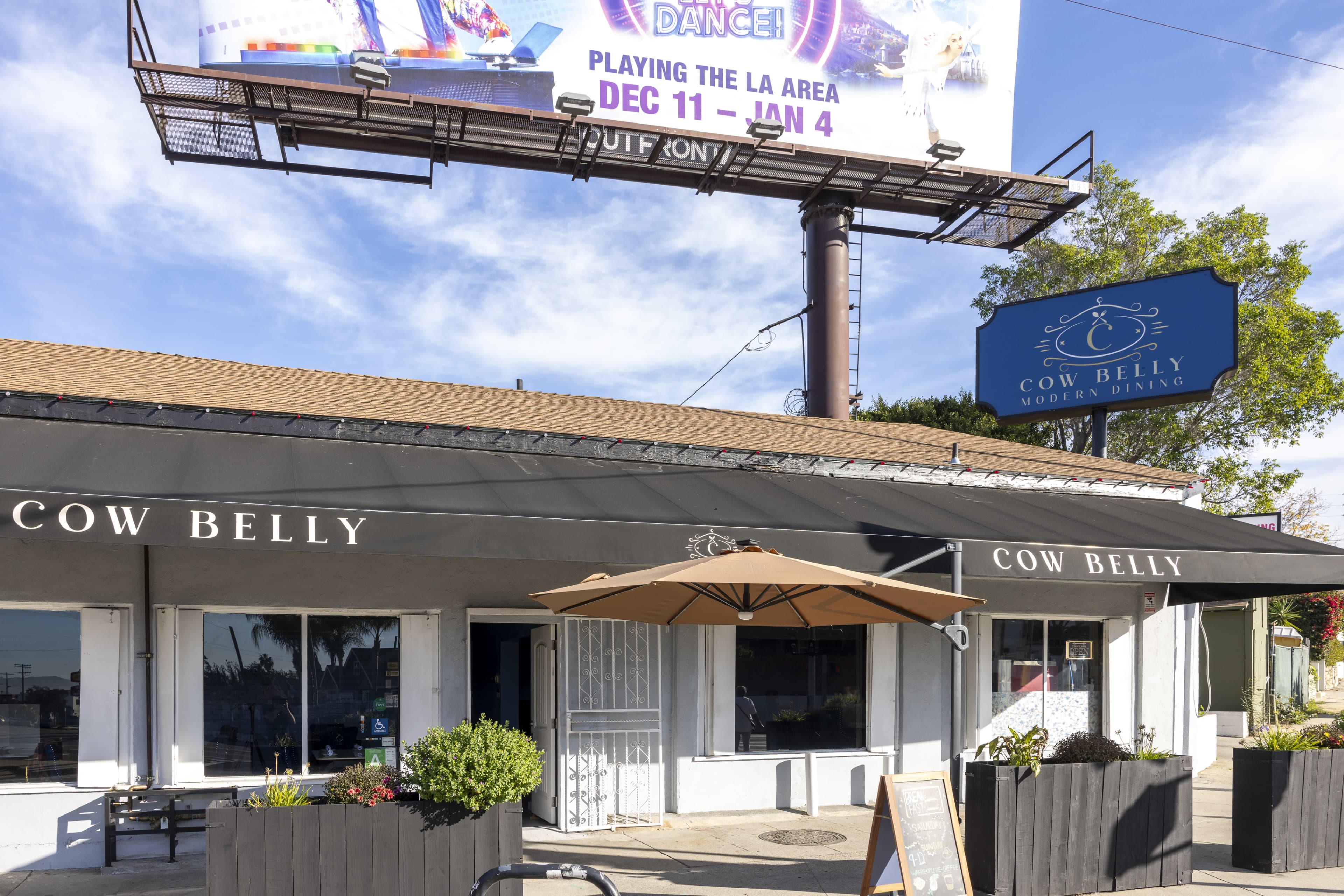 The image shows a modern dining establishment named "Cow Belly," featuring a canopy, outdoor seating, and a large billboard advertisement above.