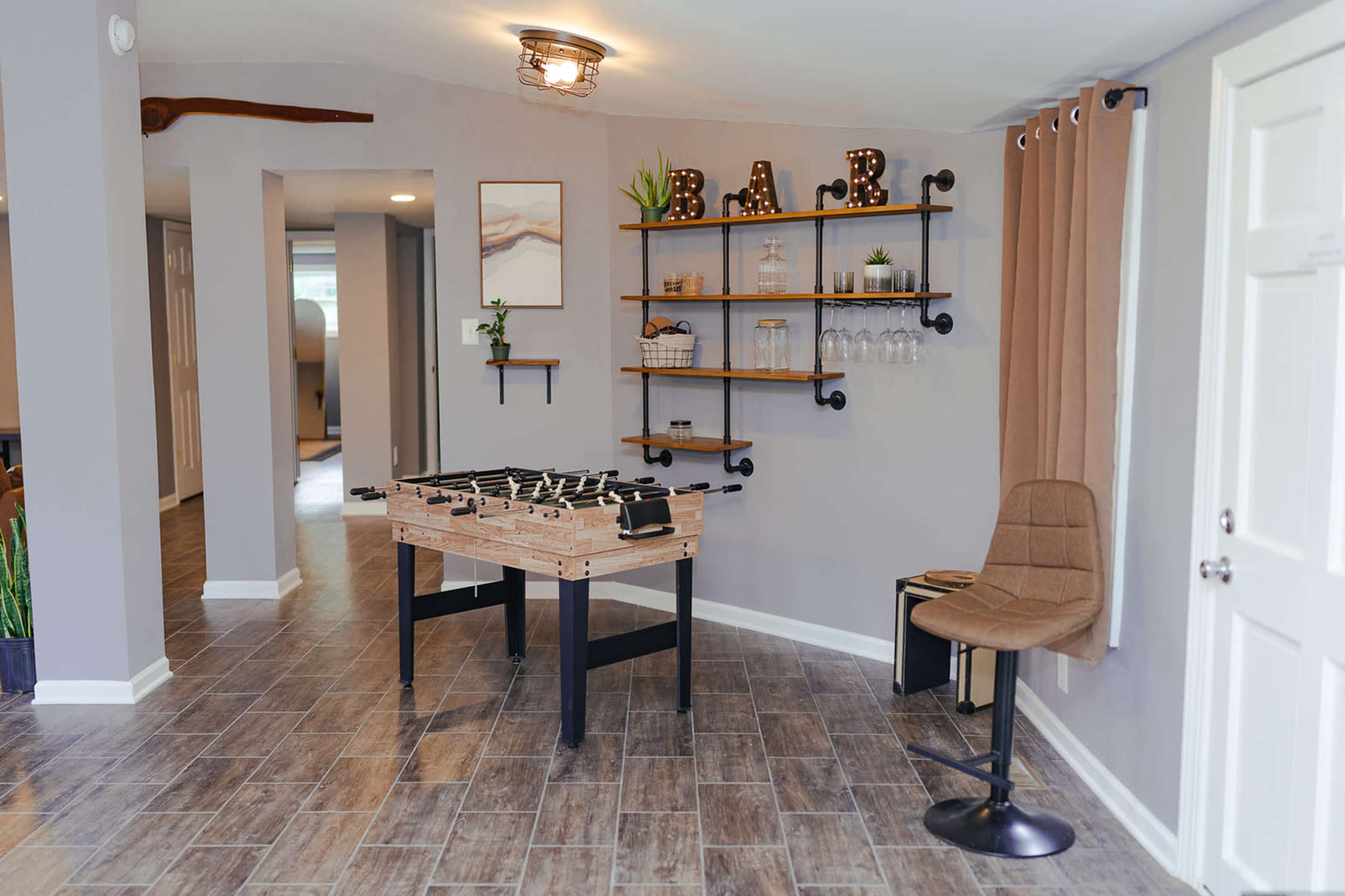 A modern, open room featuring a foosball table, a seating area with a bar stool, and industrial-style shelving displaying glassware and decorative plants.