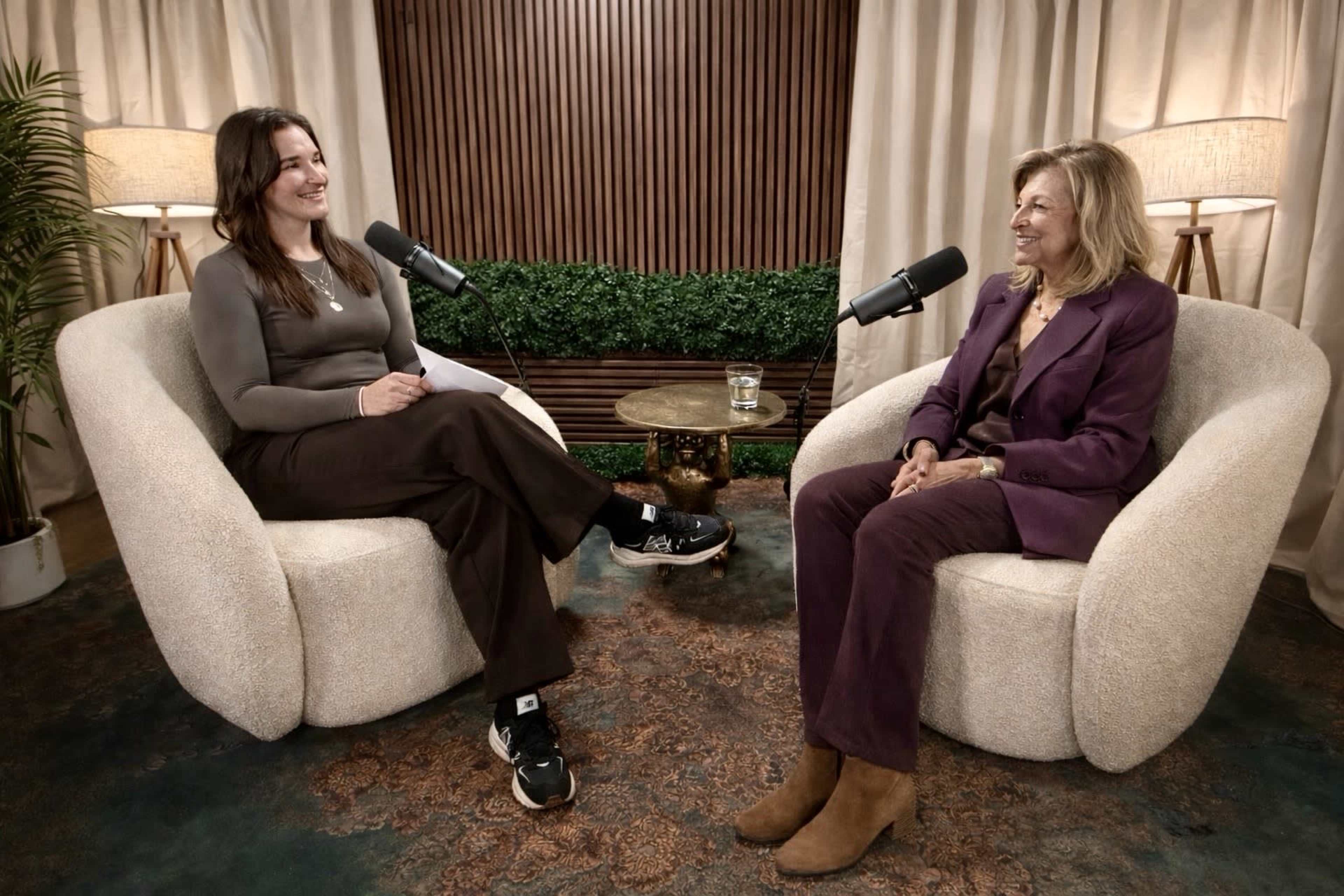 Two women sit in armchairs, speaking into microphones in a softly lit indoor setting decorated with plants and warm hues.