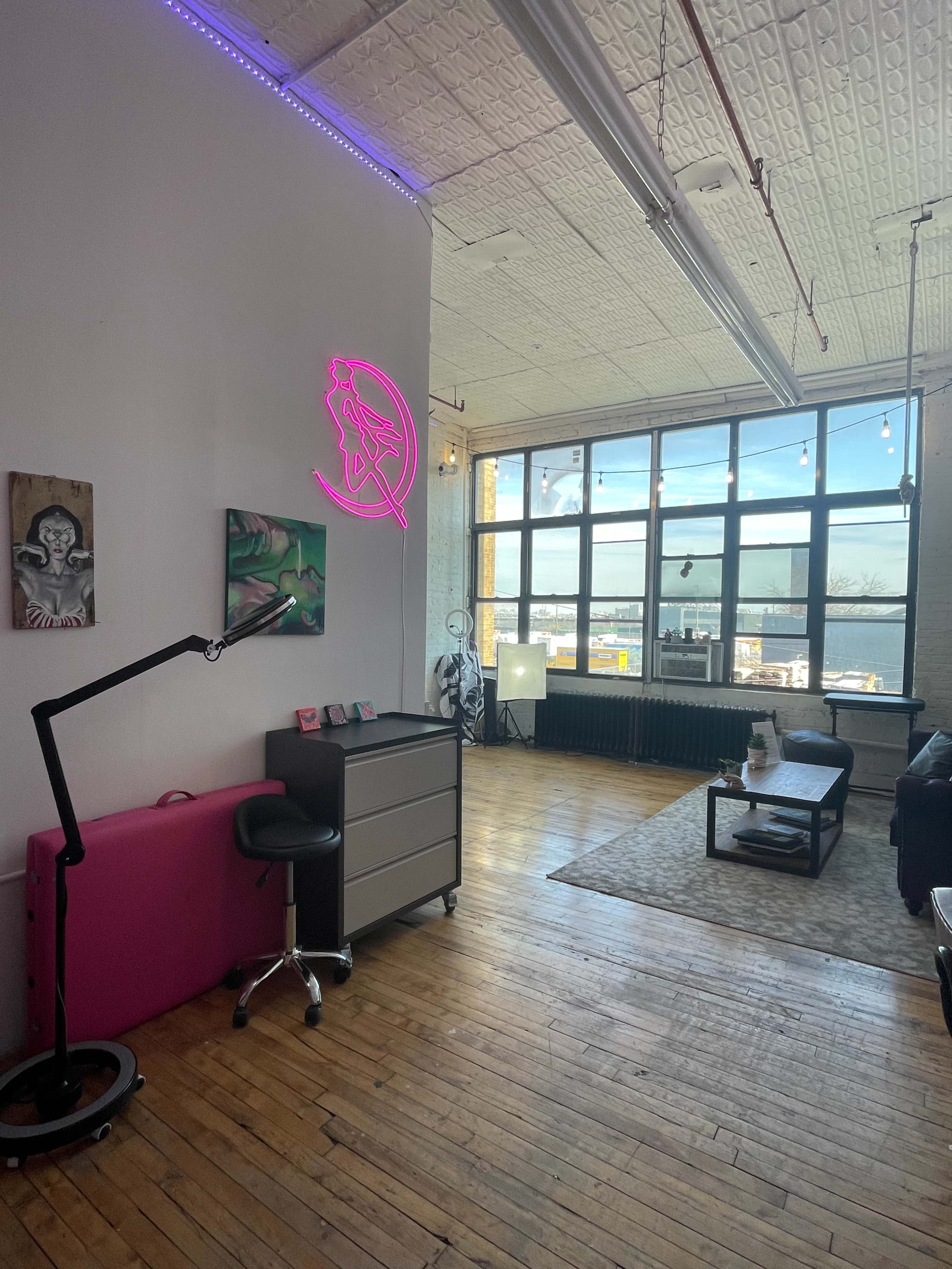 The image shows a spacious, well-lit room with large windows, wooden floors, and modern furniture, including a desk, chair, and a neon sign.