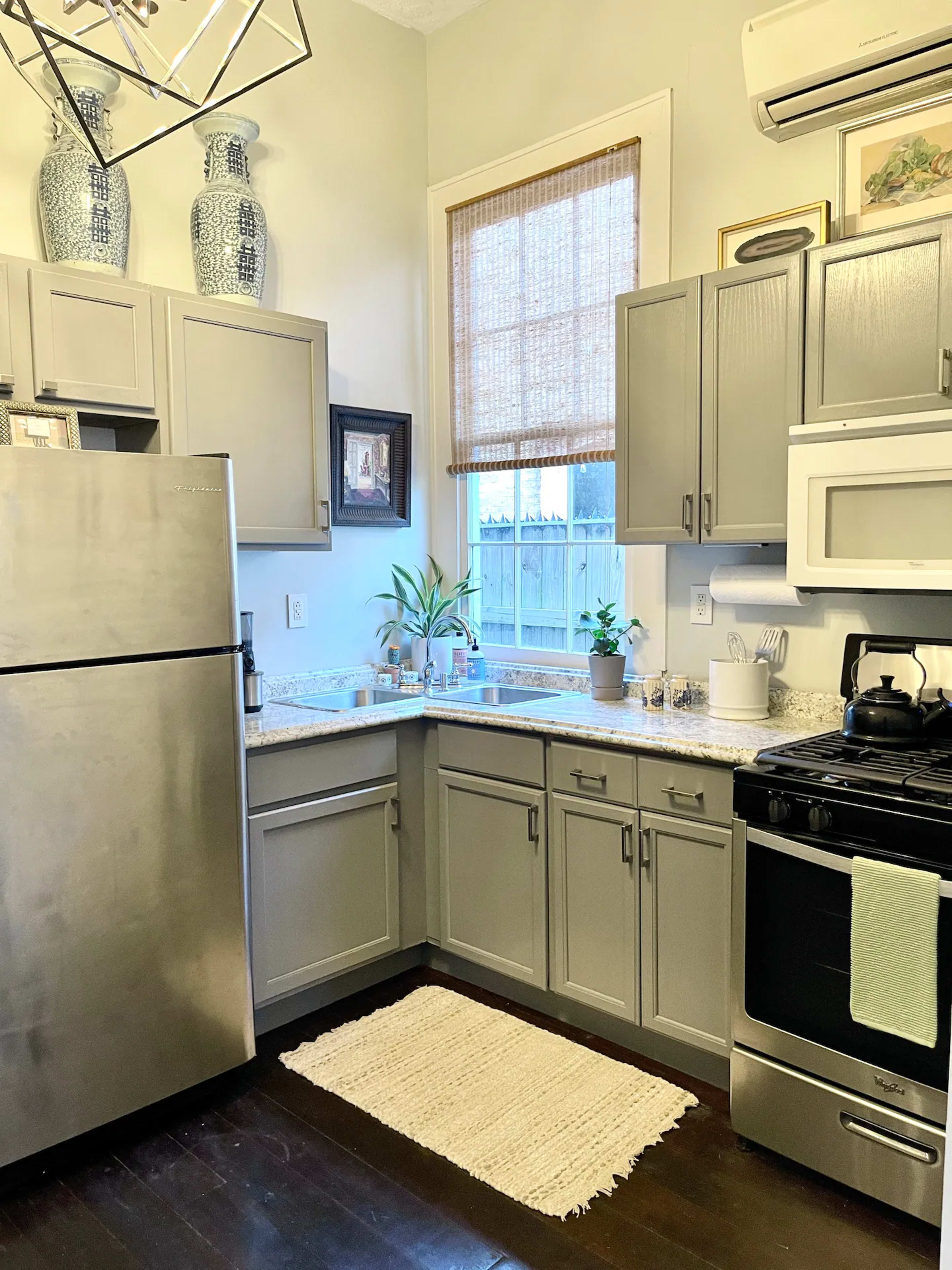 A small kitchen featuring gray cabinets, a stainless steel fridge, a gas stove, and a window with a woven shade.