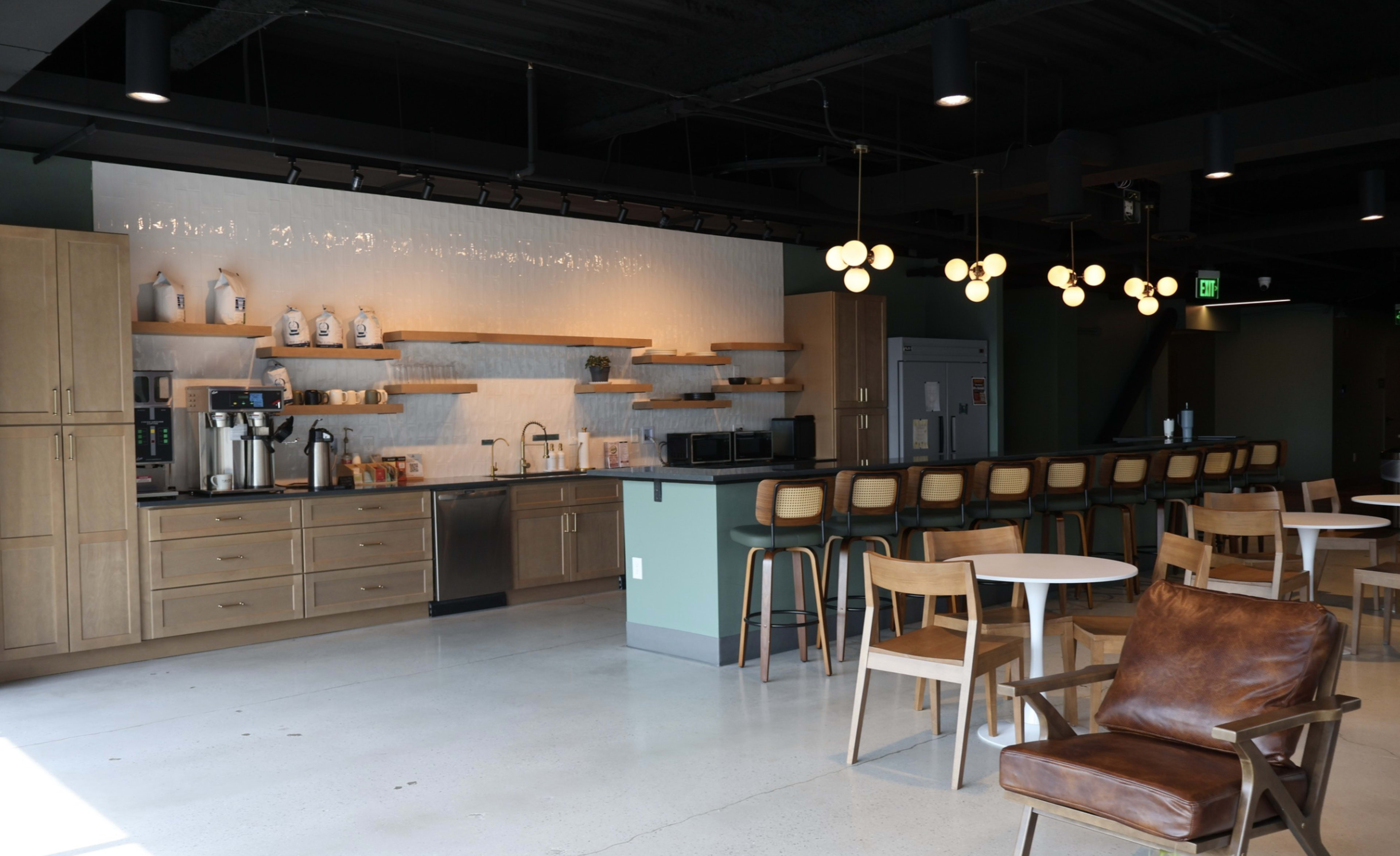 The image shows a modern cafe interior featuring a wooden countertop with various kitchen appliances, shelves, and seating arrangements including a bar with high stools and tables.