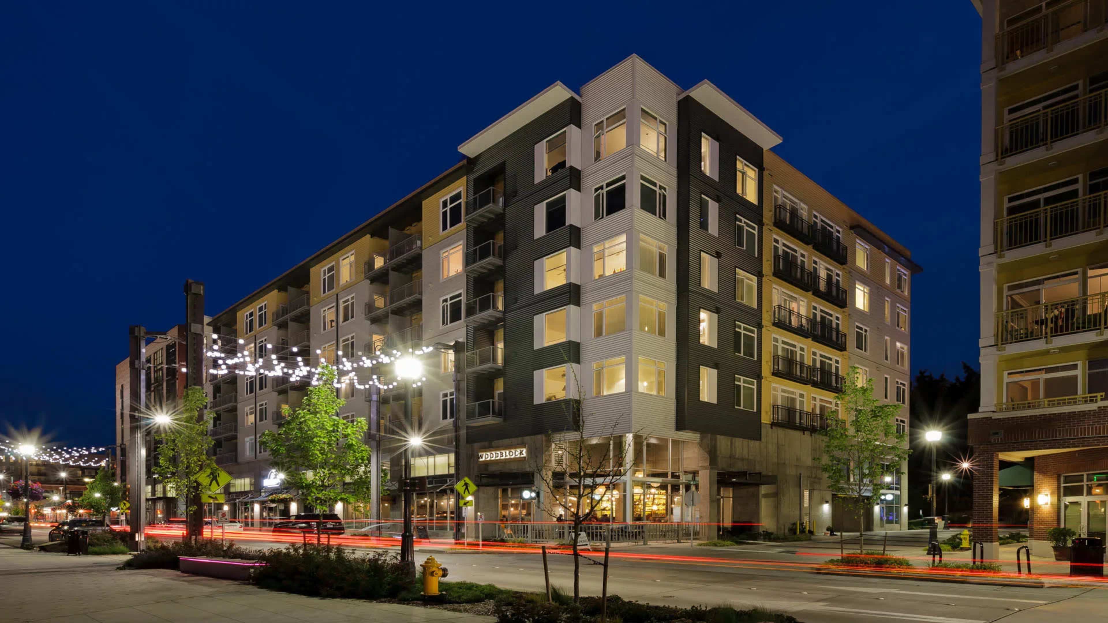 A modern multi-story apartment building at night, featuring a ground-floor storefront and streetlights illuminating the surrounding area.