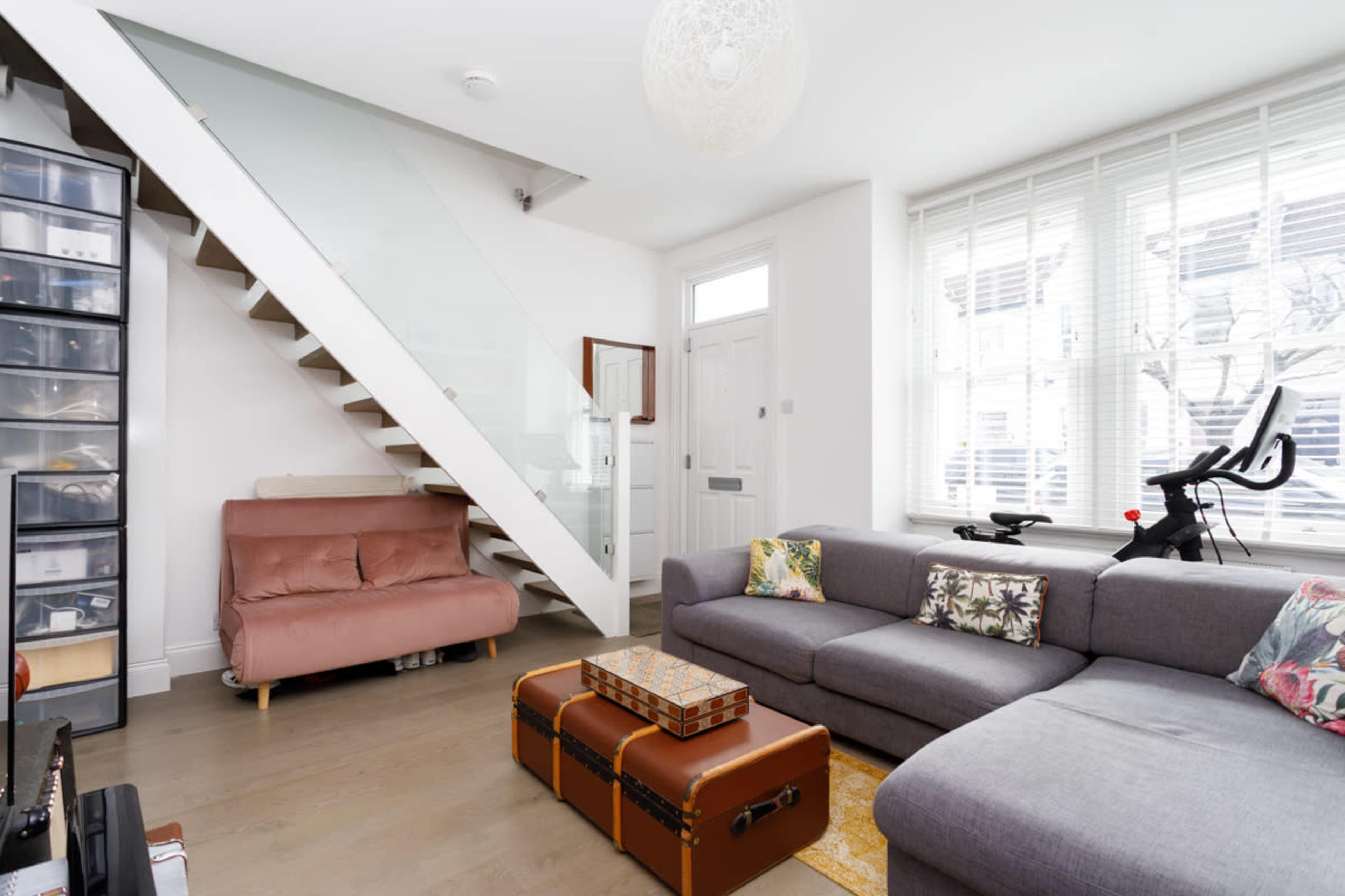 The image shows a modern living room with a gray sectional sofa, a pink couch, a small coffee table, and a staircase that leads to an upper level.