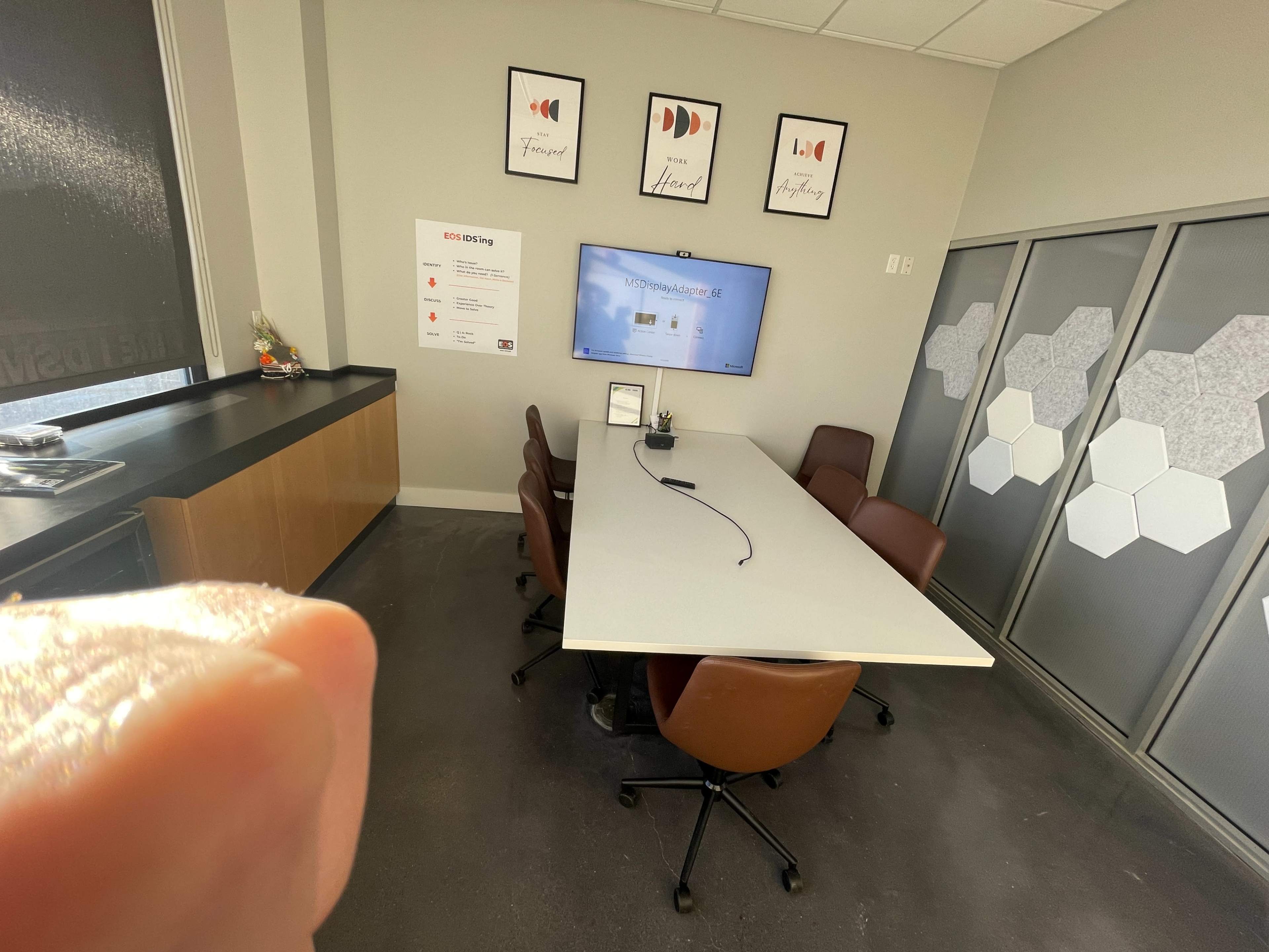 The image shows a conference room with a long table surrounded by brown chairs, a large screen displaying information, and decorative framed prints on the wall.