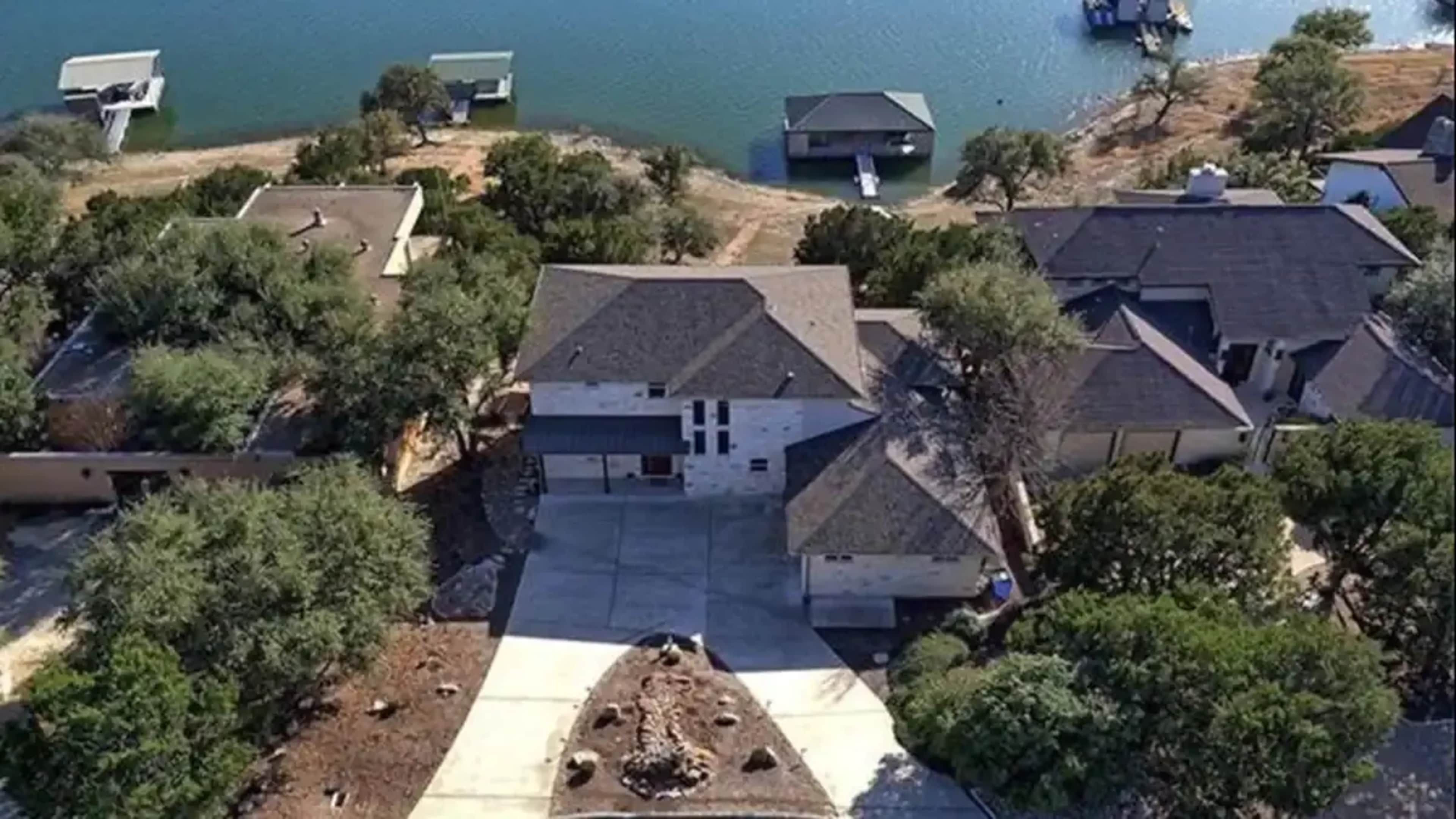 An aerial view of a house situated near a body of water, with several nearby structures and trees.