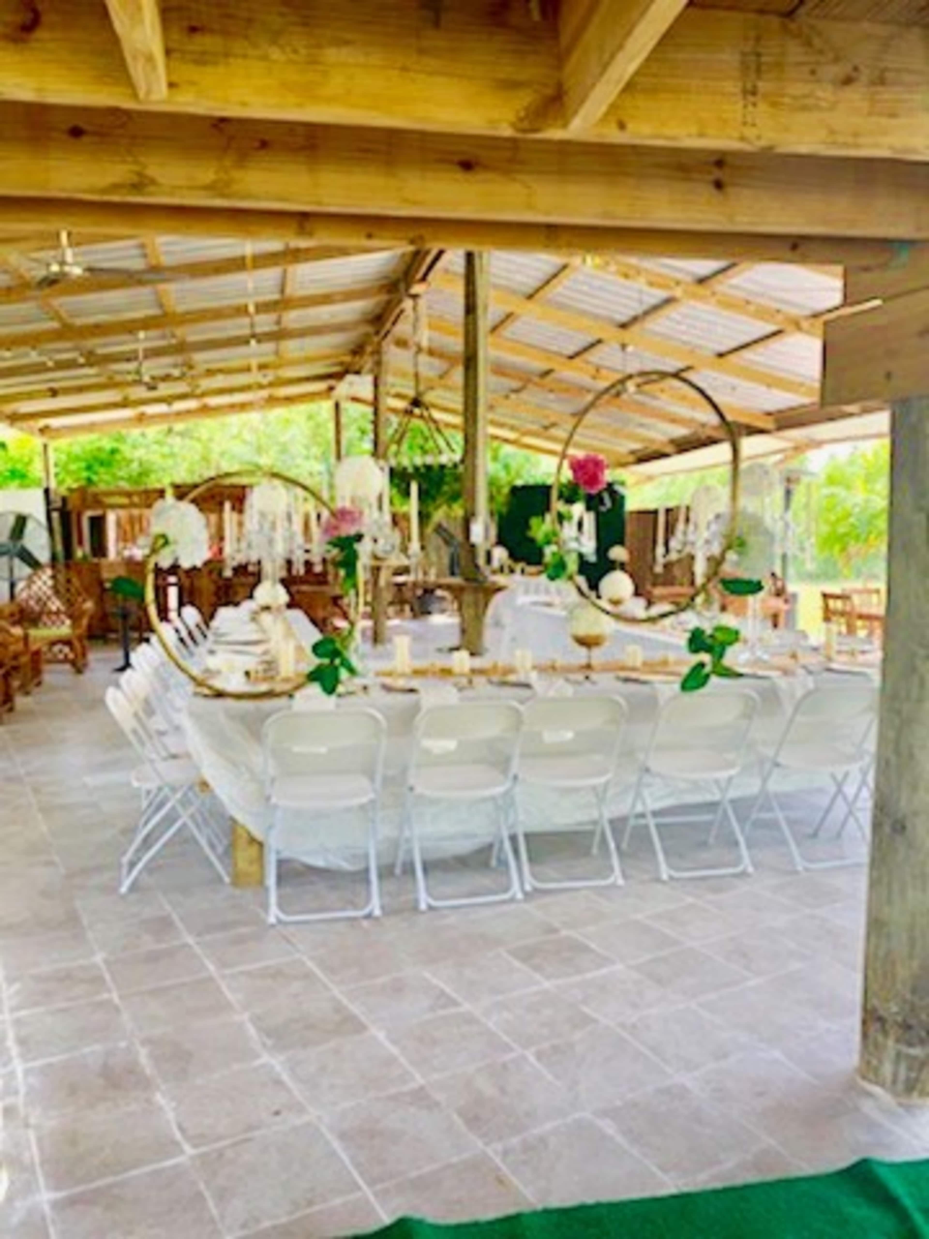 A long, elegantly set dining table with white chairs is arranged under a wooden pavilion, surrounded by greenery.