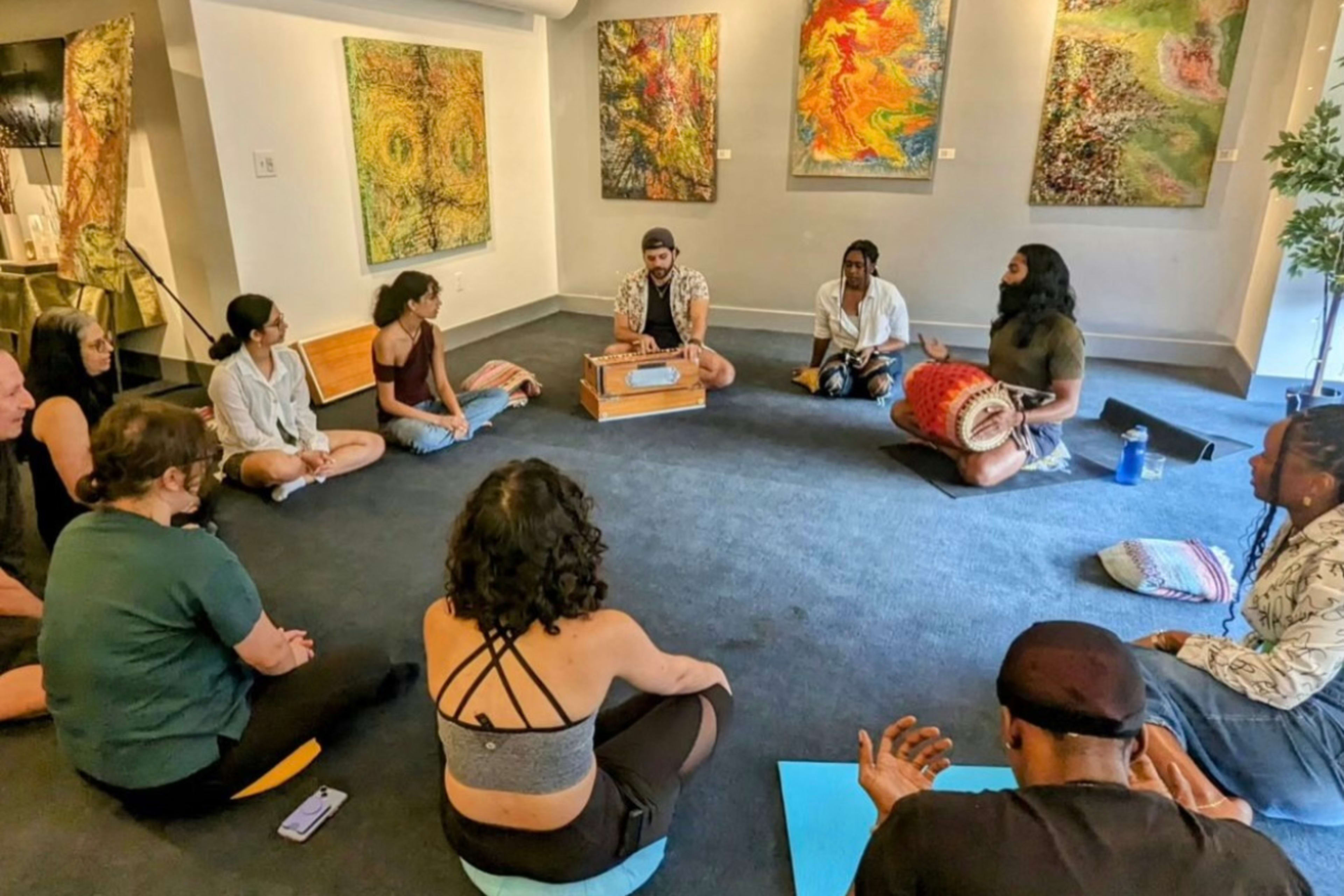 A group of people sit in a circle on the floor of an art gallery, engaging in an interactive session while surrounded by colorful abstract paintings on the walls.