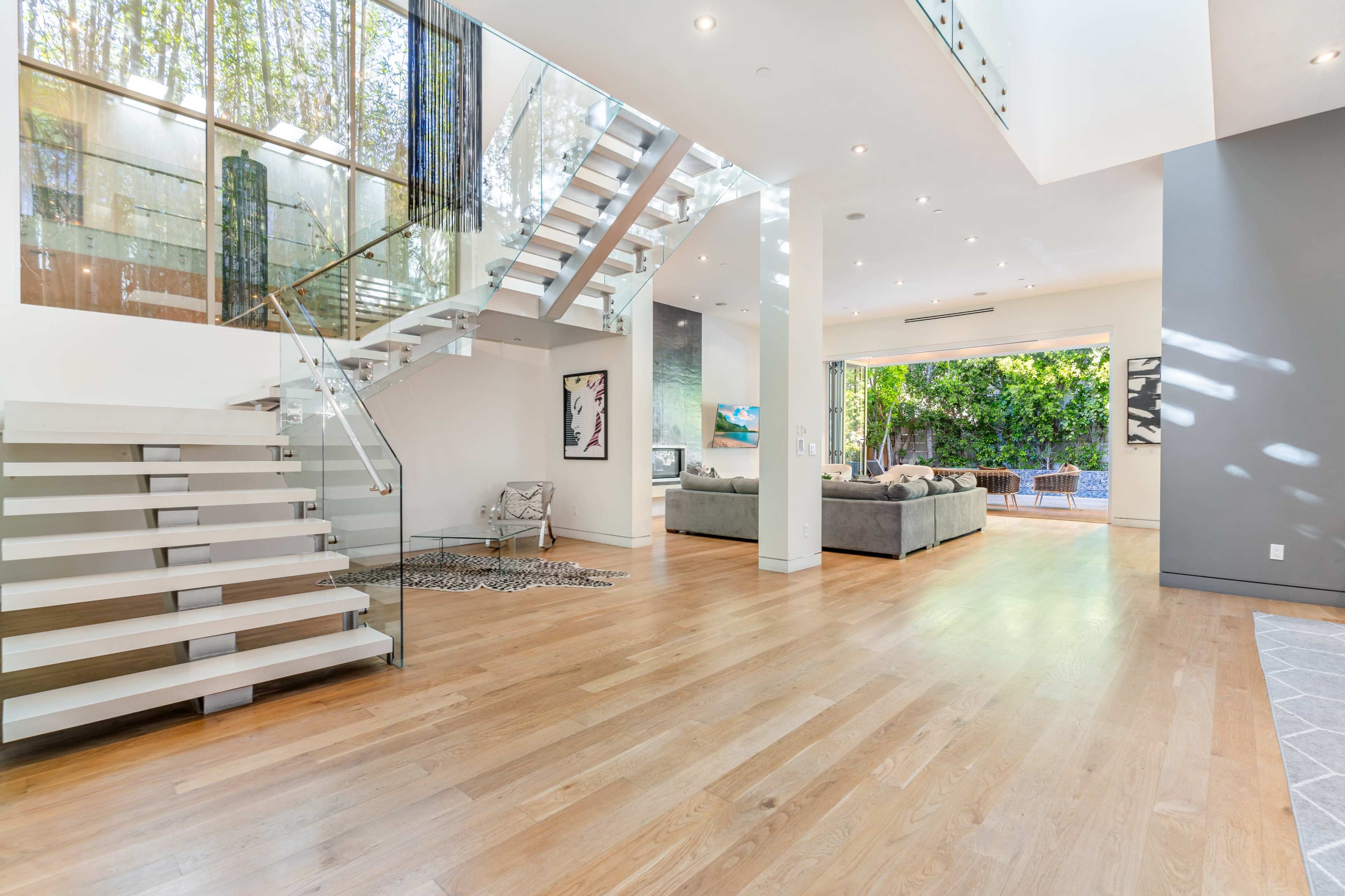 A spacious, modern living area featuring a glass staircase, large windows, and sliding doors opening to a lush outdoor space.
