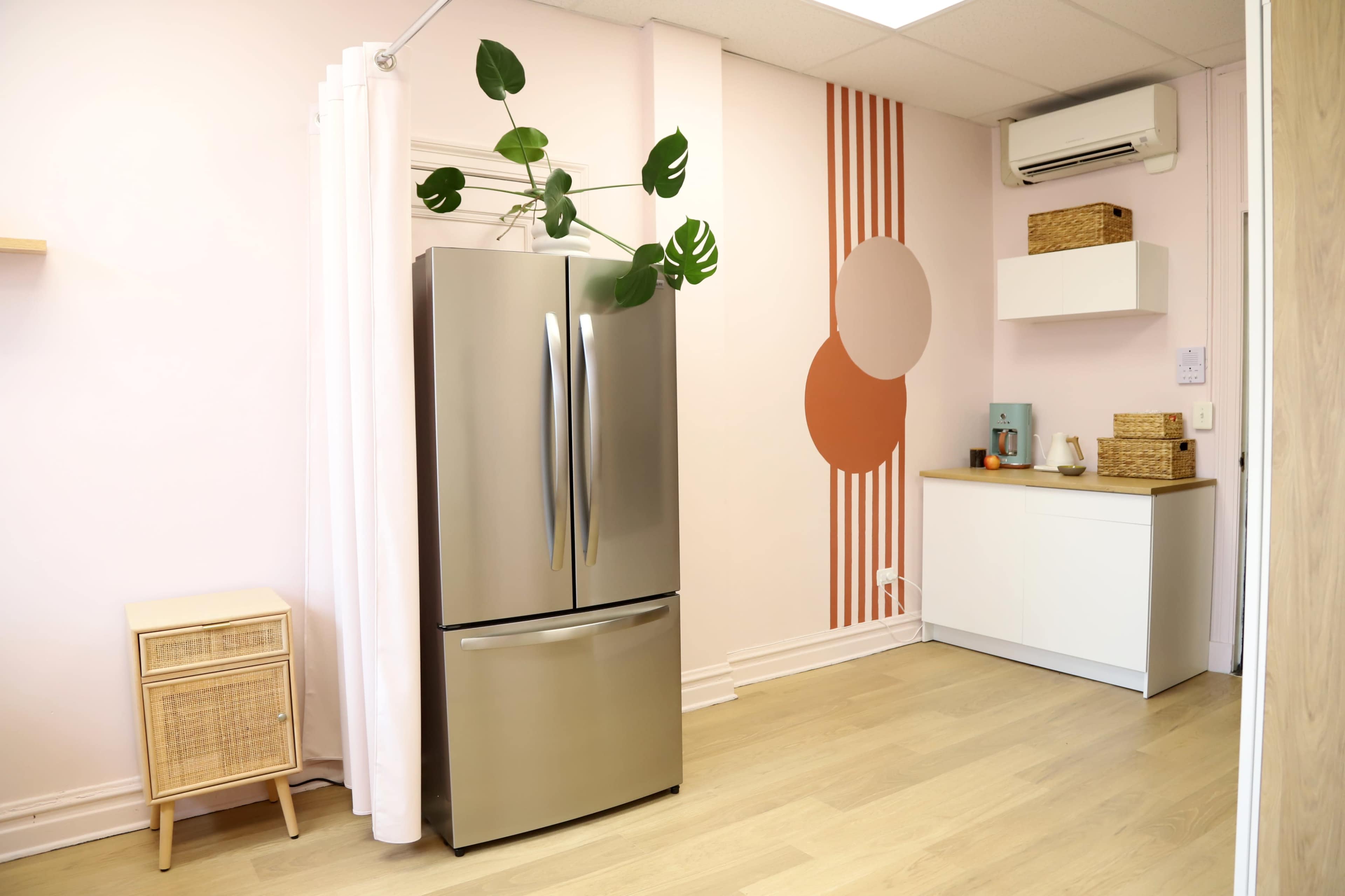 The image shows a modern, minimalistic kitchen space featuring a stainless steel refrigerator, a white cabinet, and decorative wall elements in pastel colors.