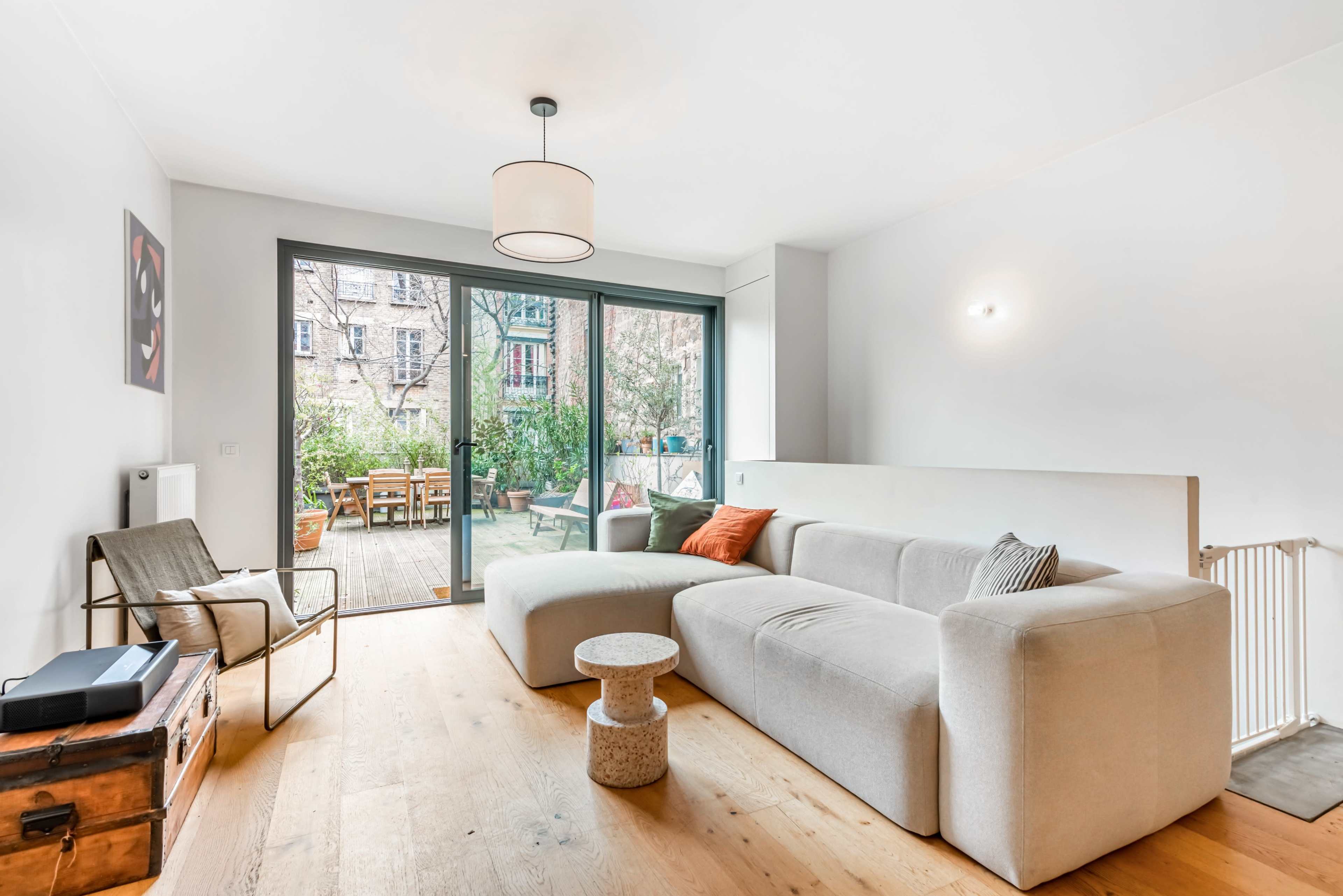 A modern living room with a grey sectional sofa, a round side table, and large glass doors opening to a wooden deck with outdoor furniture.