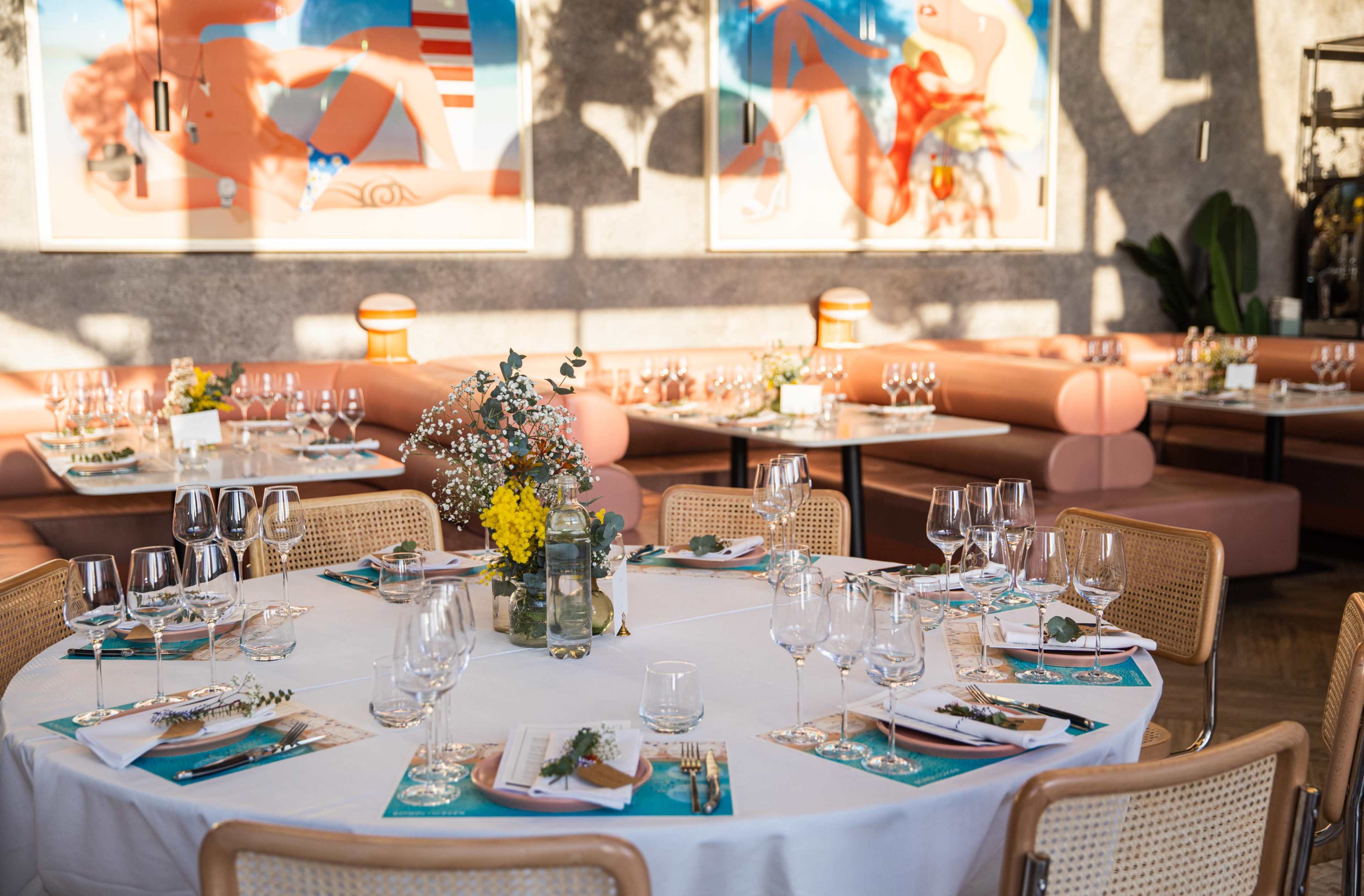 A dining table is elegantly set with glassware and decorative flowers, surrounded by comfortable seating in a brightly lit restaurant adorned with modern artwork.