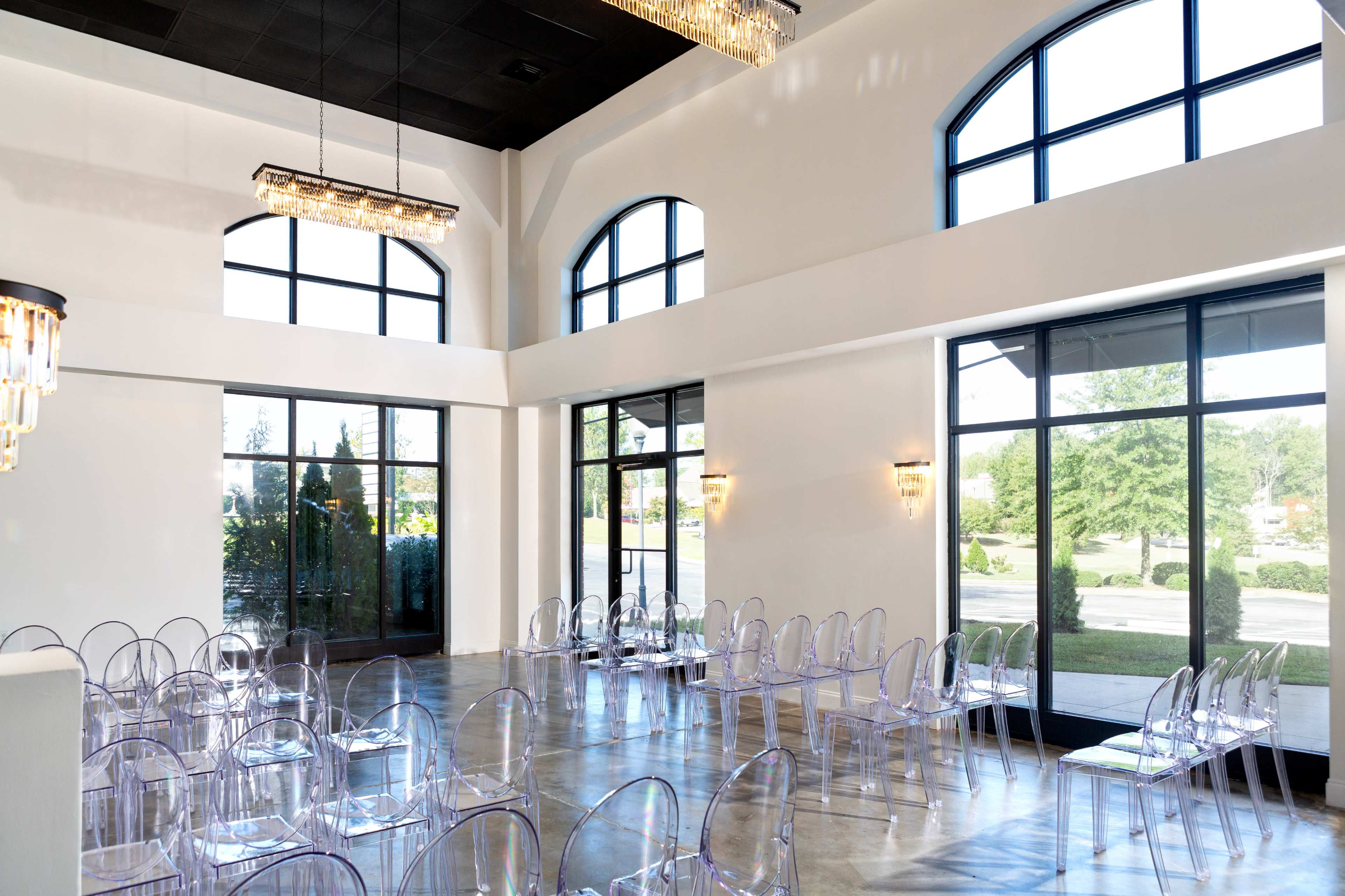 A spacious event room features clear acrylic chairs arranged neatly, large windows allowing natural light, and modern chandeliers above.