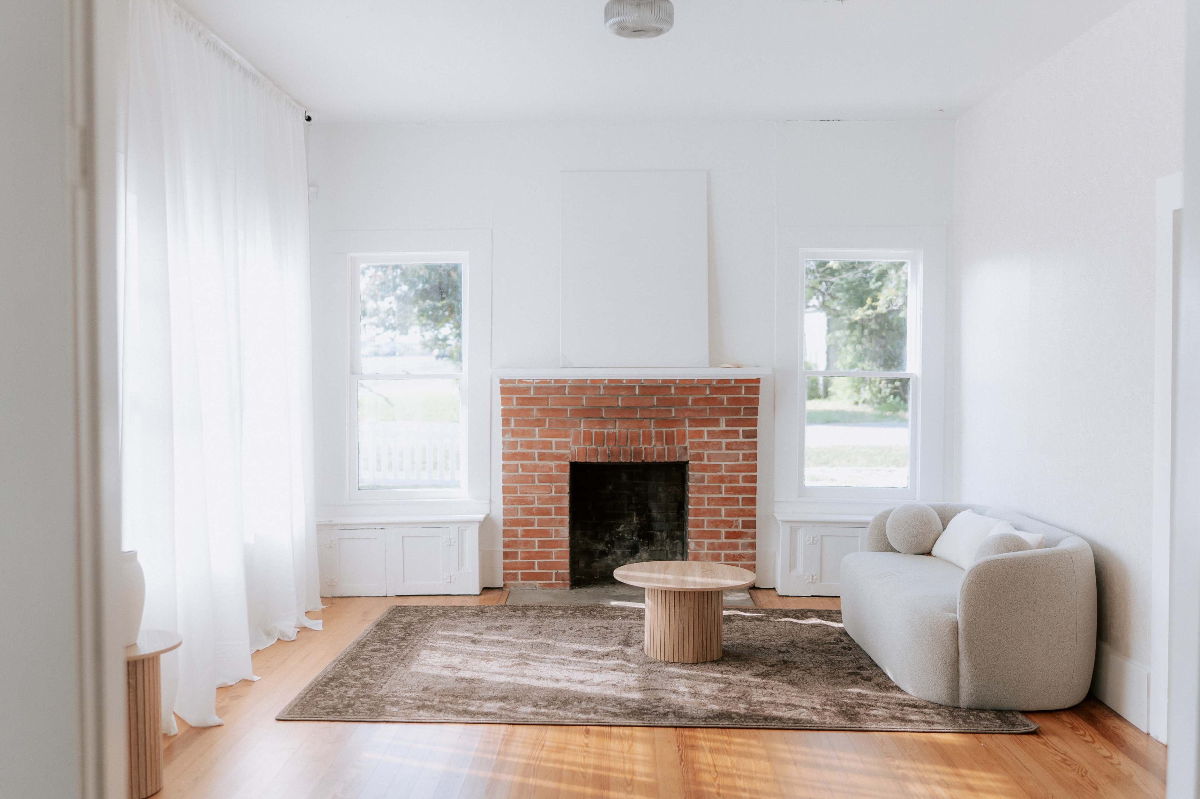 A minimalist living room features a brick fireplace, a round coffee table, a light-colored sofa, and large windows with sheer curtains.
