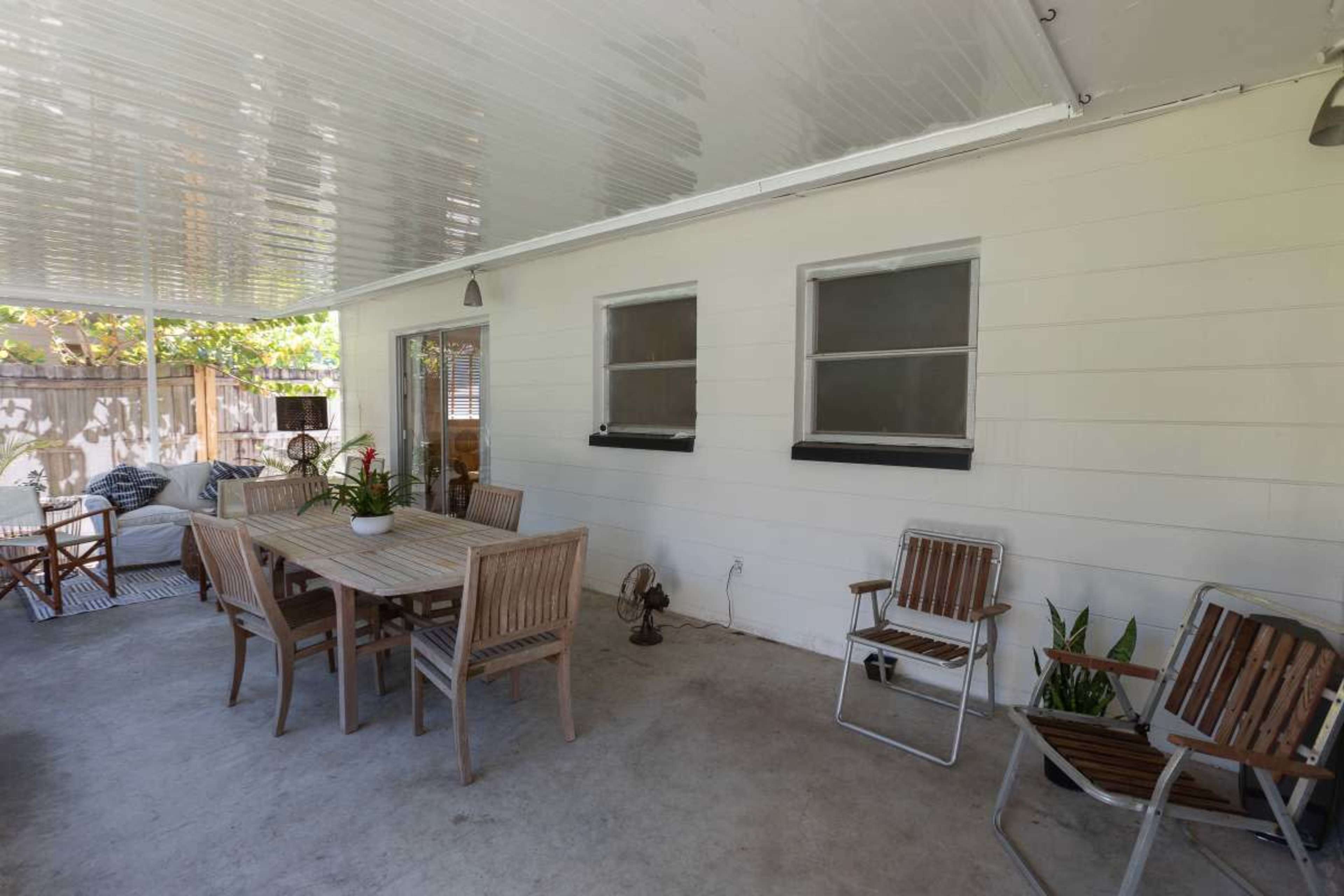 The image shows a covered outdoor patio area with a wooden dining table, chairs, and potted plants against a white wall.