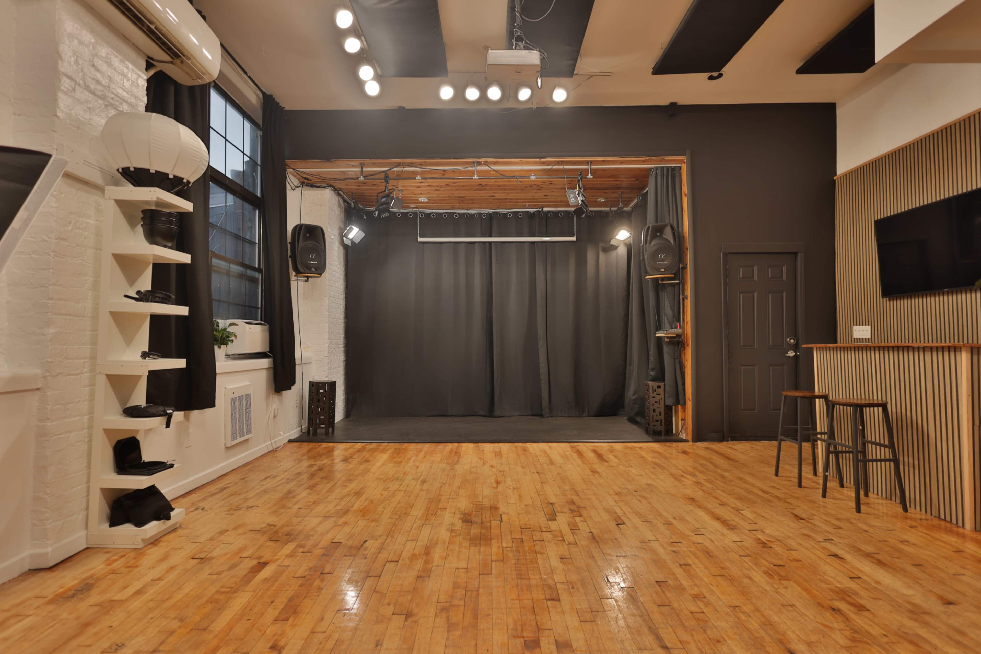 The image shows a spacious, well-lit studio with wooden floors, a black curtain backdrop, and various stage lighting fixtures.